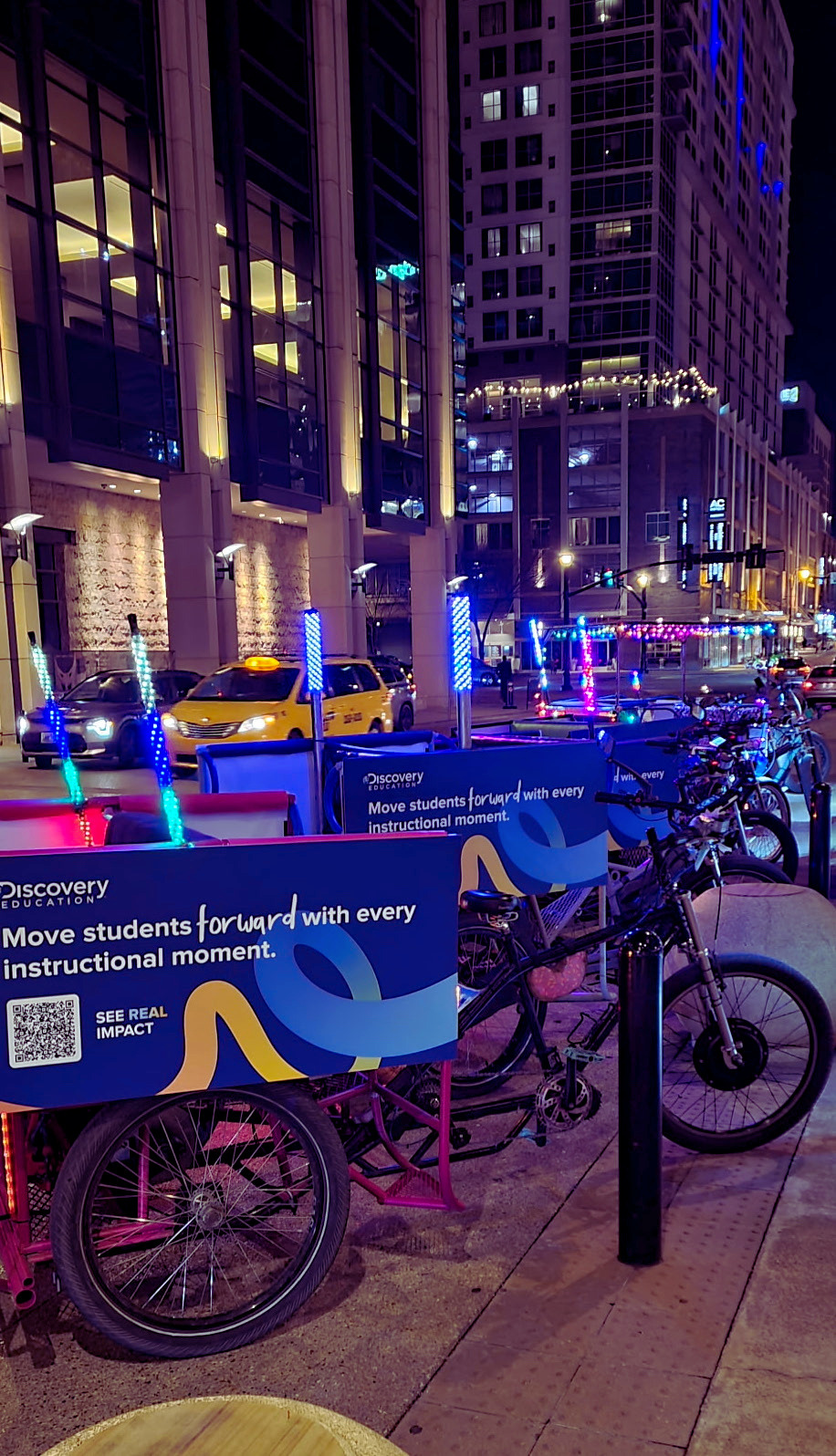 Nighttime city street scene with illuminated signs, parked bikes with Discovery Education signs, taxis, and tall modern buildings.