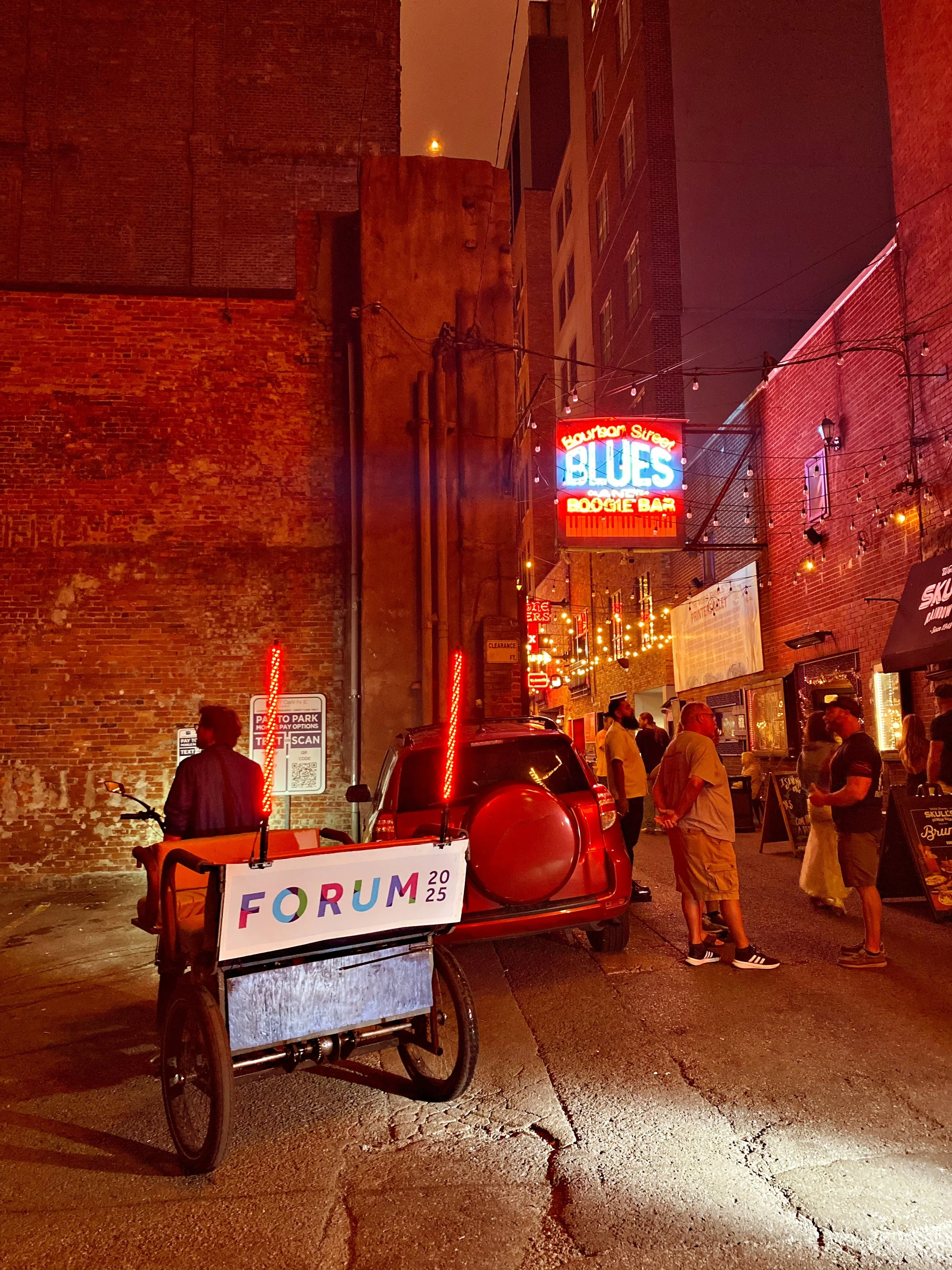 Night scene on Beale Street in Memphis showing a neon sign for Blues on Beale Street, a small group of people talking, a bicycle with a sign reading 'Forum 2025', parked cars, and string lights.