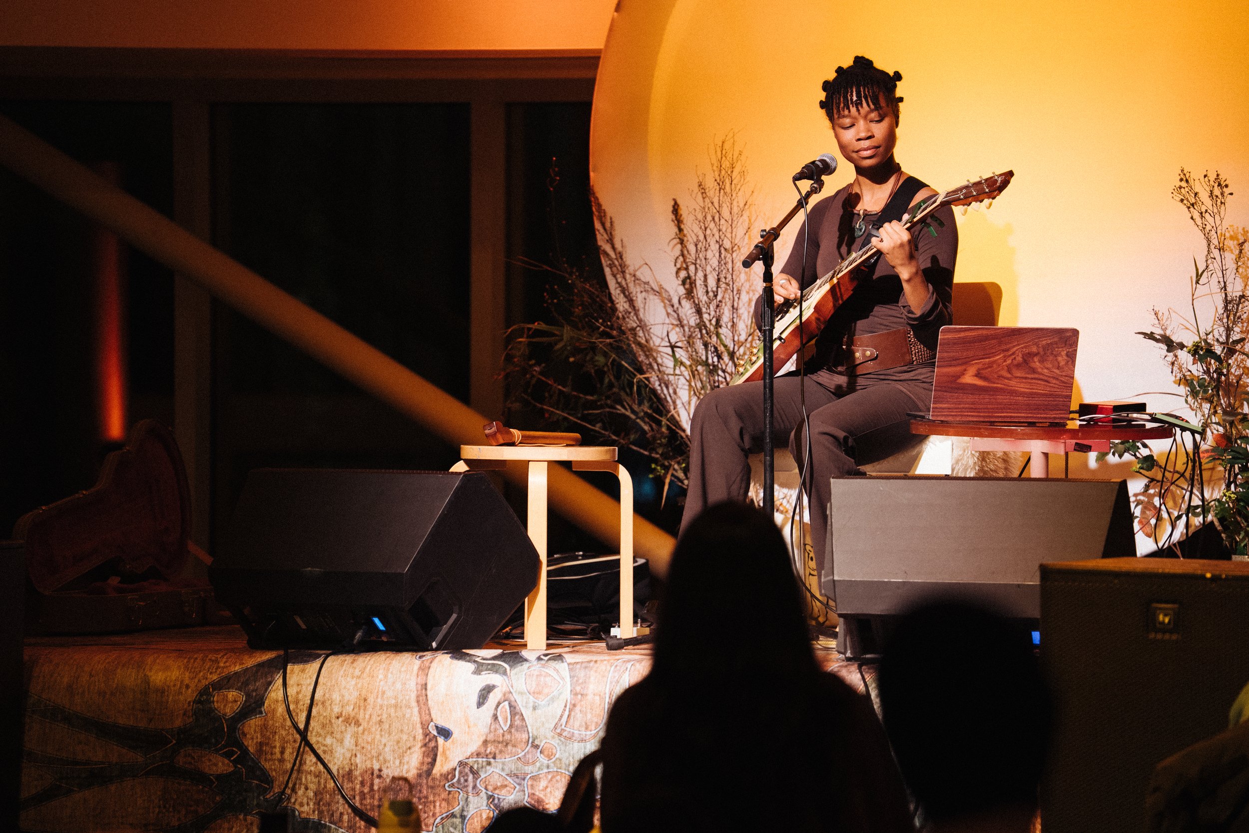 Artist Dianna Lopez sitting on a stage looking at their guitar with grasses and an orange circle background behind them.