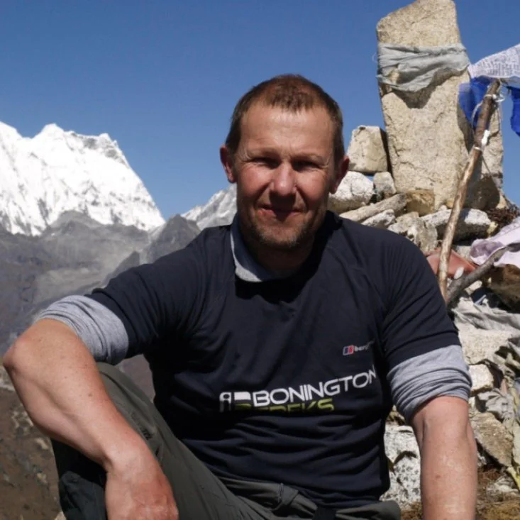 Joe Bonington sitting on rocky terrain during a mountain expedition, with snow-capped mountains in the background and prayer flags nearby.