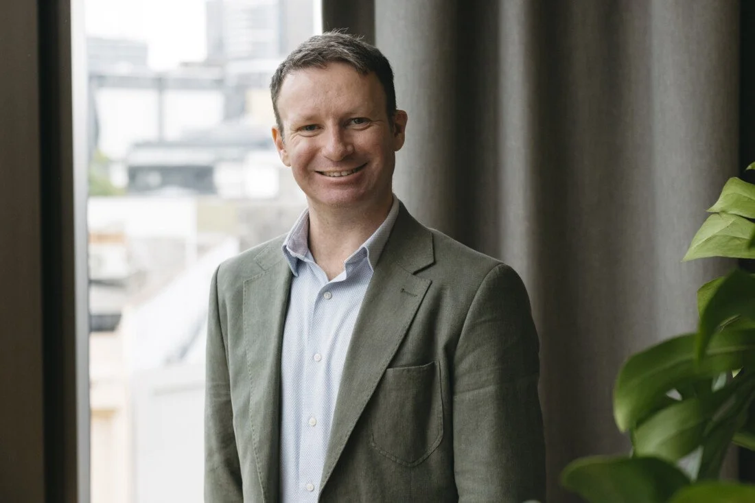 Portrait of a smiling man in a gray blazer and light blue shirt standing near a window with city buildings in the background.