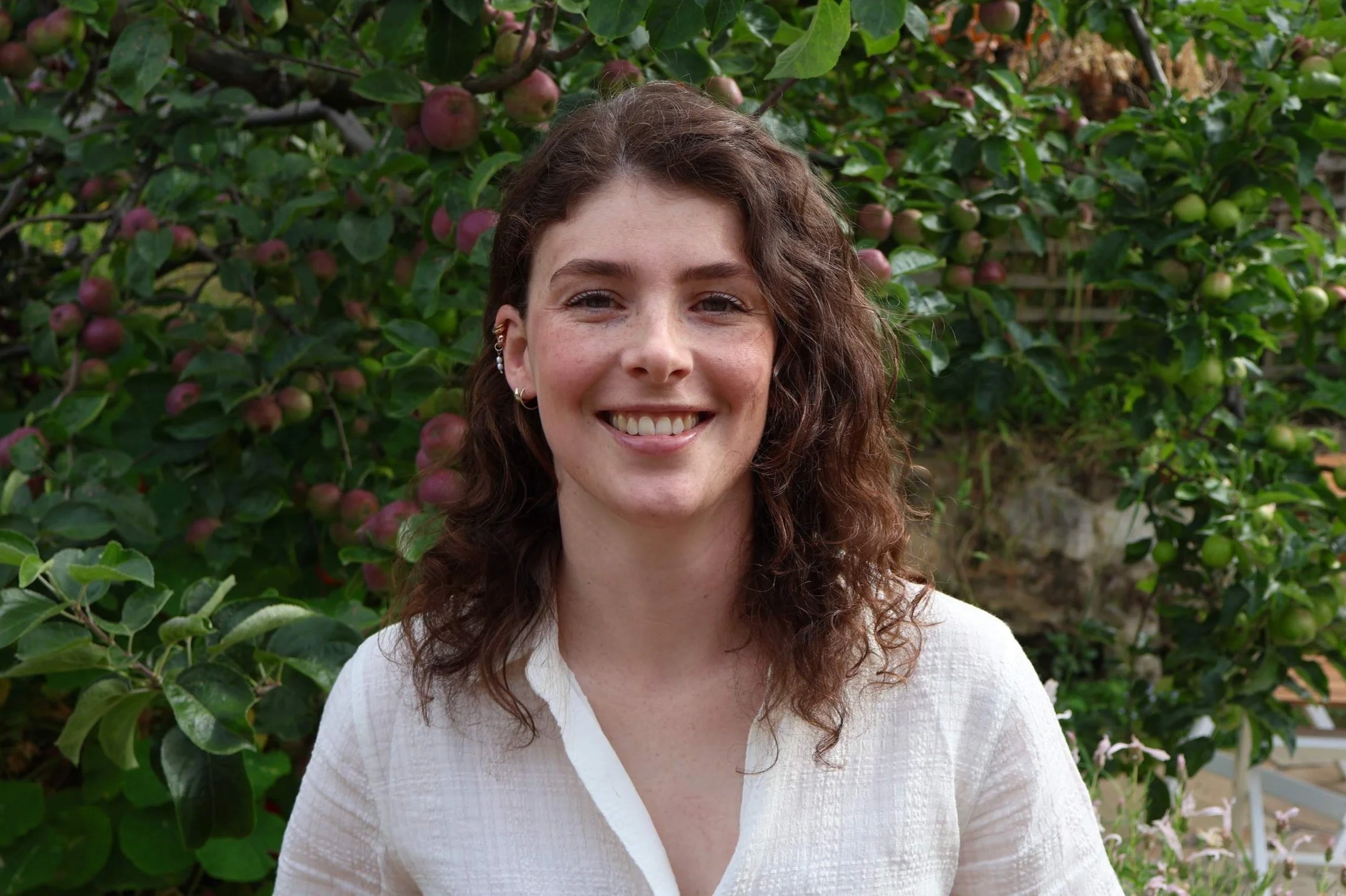 A woman with curly brown hair smiling, standing in front of an apple tree with ripe and green apples.