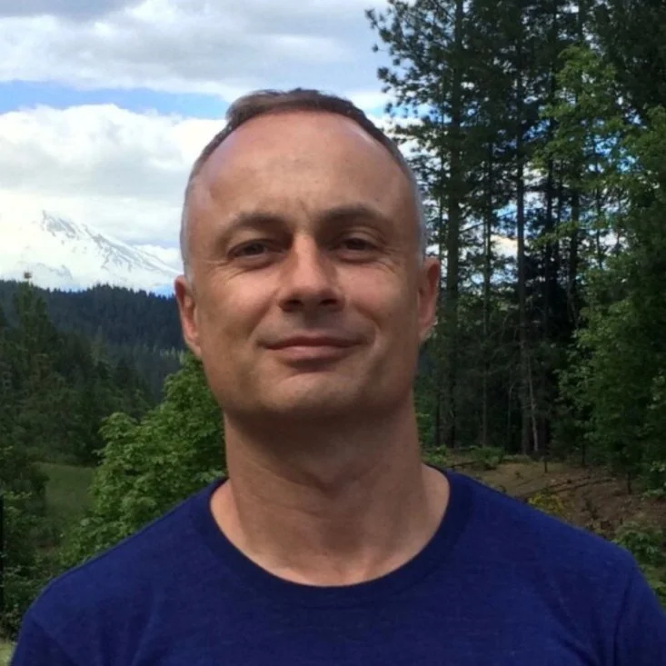 A man standing outdoors in front of a forested landscape with a snowy mountain in the background.