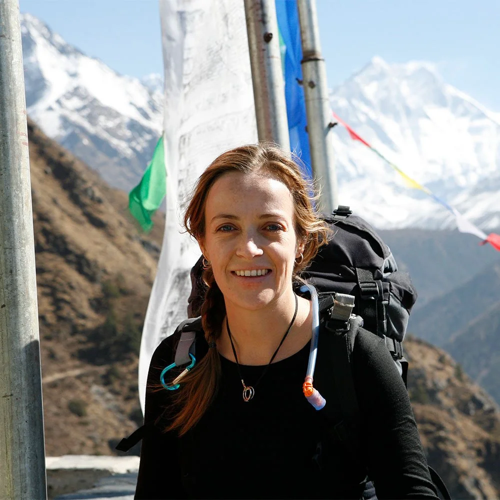 Jen Peedom with auburn hair smiling, wearing a black shirt and carrying a large backpack, standing outdoors with snow-capped mountains in the background.