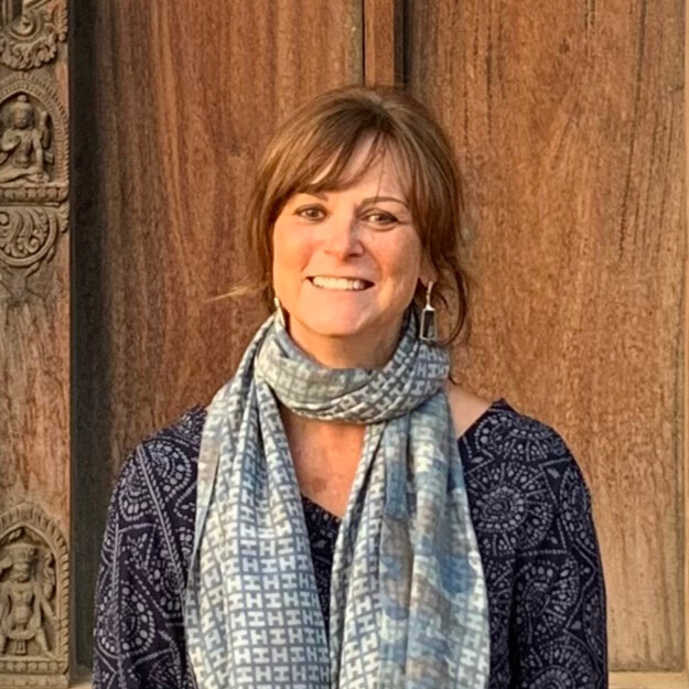 A woman with short brown hair smiling at the camera, wearing a dark patterned top and a blue patterned scarf, standing in front of a wooden background with carved details.