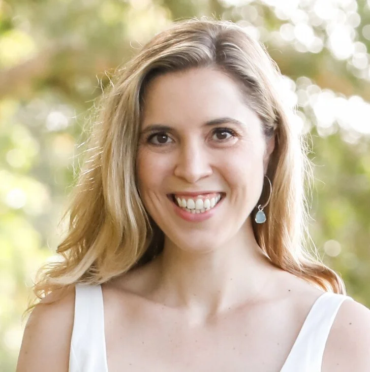 A woman with long, wavy blonde hair smiling outdoors, wearing a white sleeveless top and a dangling earring, with a blurred background of trees and sunlight.