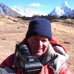 Michael Dillon in outdoor gear holding camera with mountains and snow in the background.