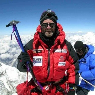 A man dressed in red mountaineering gear smiling on a snowy mountain with other climbers and ice-covered peaks in the background.