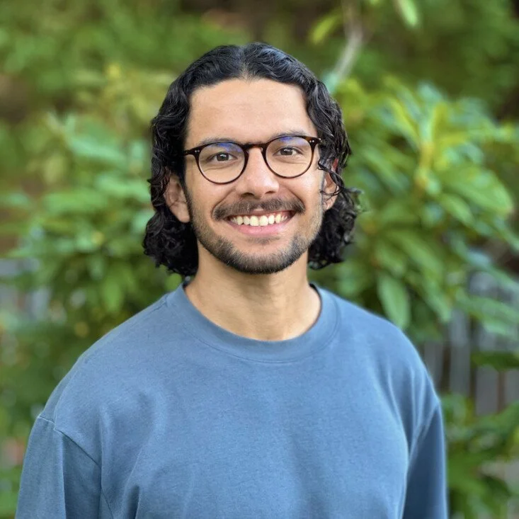 A young man with curly dark hair and glasses smiling outdoors with greenery in the background.