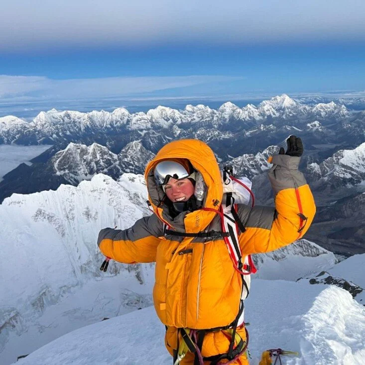 Gabby Kanizay in yellow and gray mountaineering gear smiling and waving at the summit of Mount Everest with others in the background and a clear blue sky.