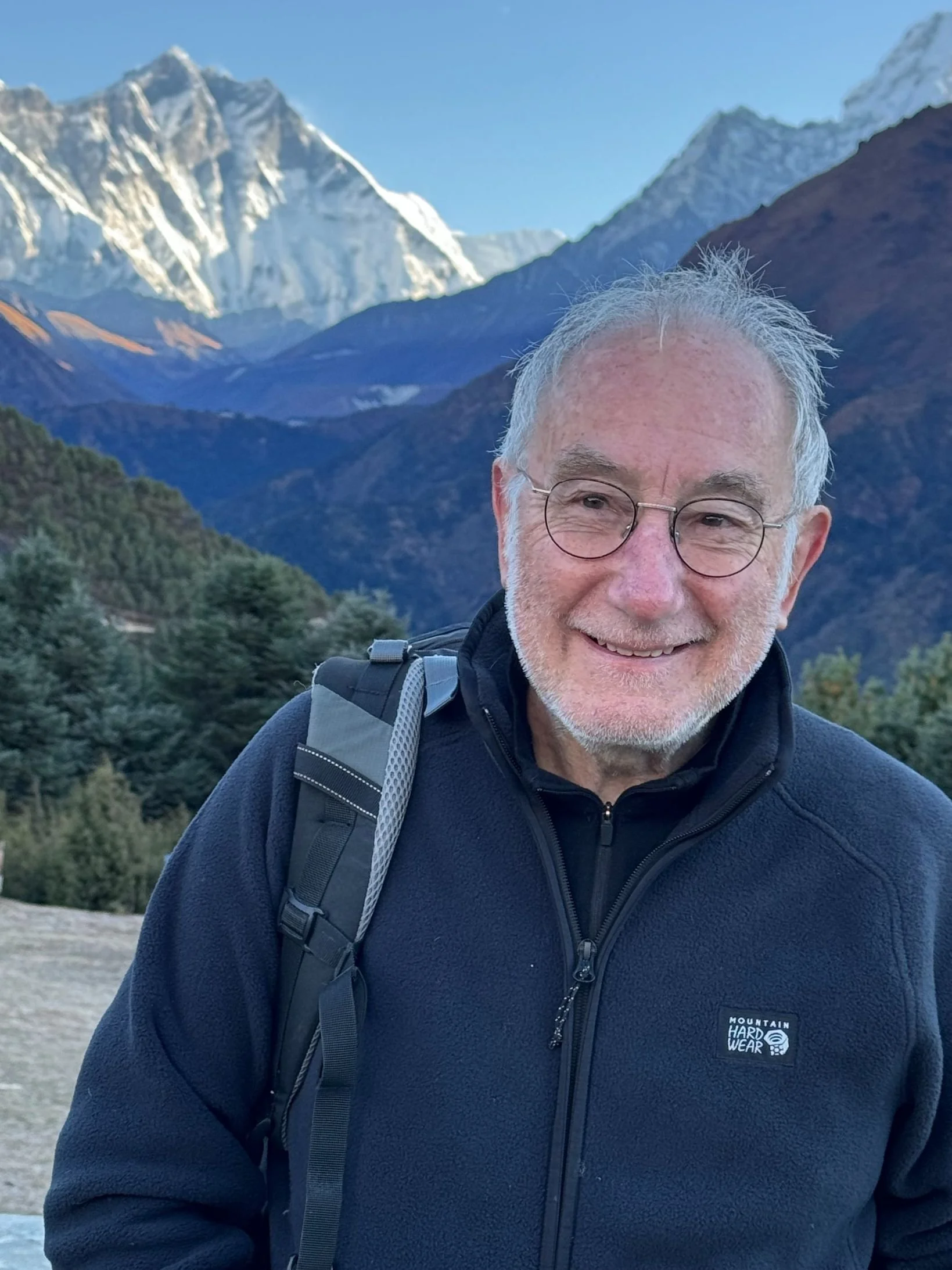 An elderly man with glasses and a backpack smiling in front of snow-capped mountains and green forested hills.