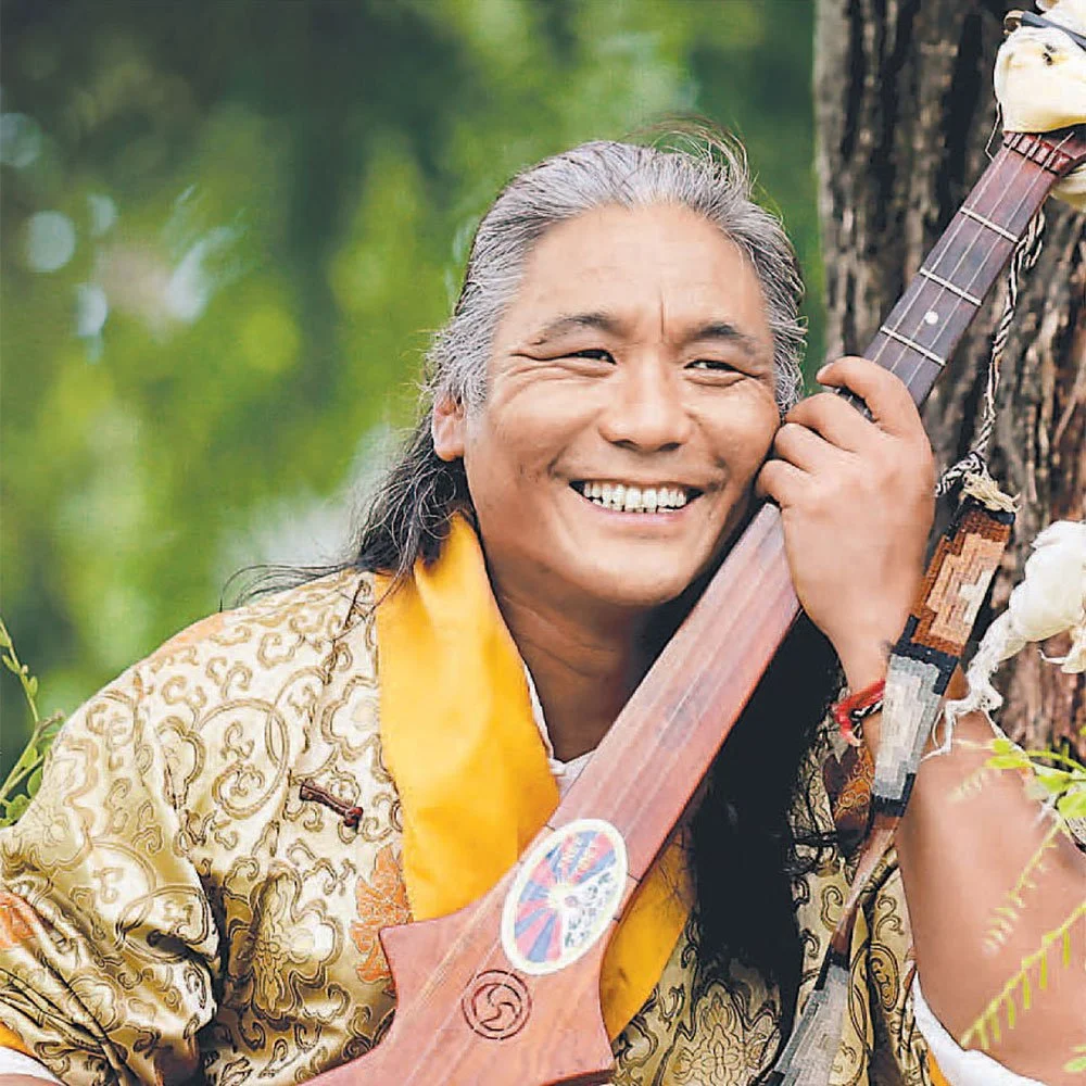 Tenzin ChoegyalA smiling man with gray hair in a patterned jacket is holding a stringed musical instrument close to his face. He is outdoors with a green, blurred background.