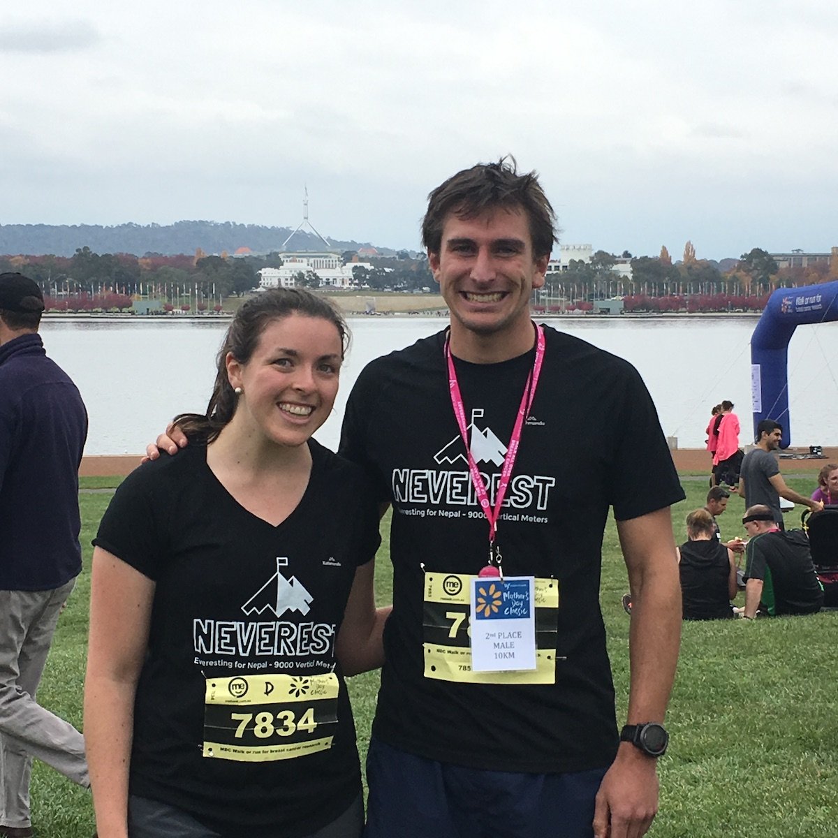 Two smiling runners, woman and man, wearing black 'NEVERREST' T-shirts and race bibs, standing outdoors near a lake with people in the background, likely after participating in a race event in an urban park.