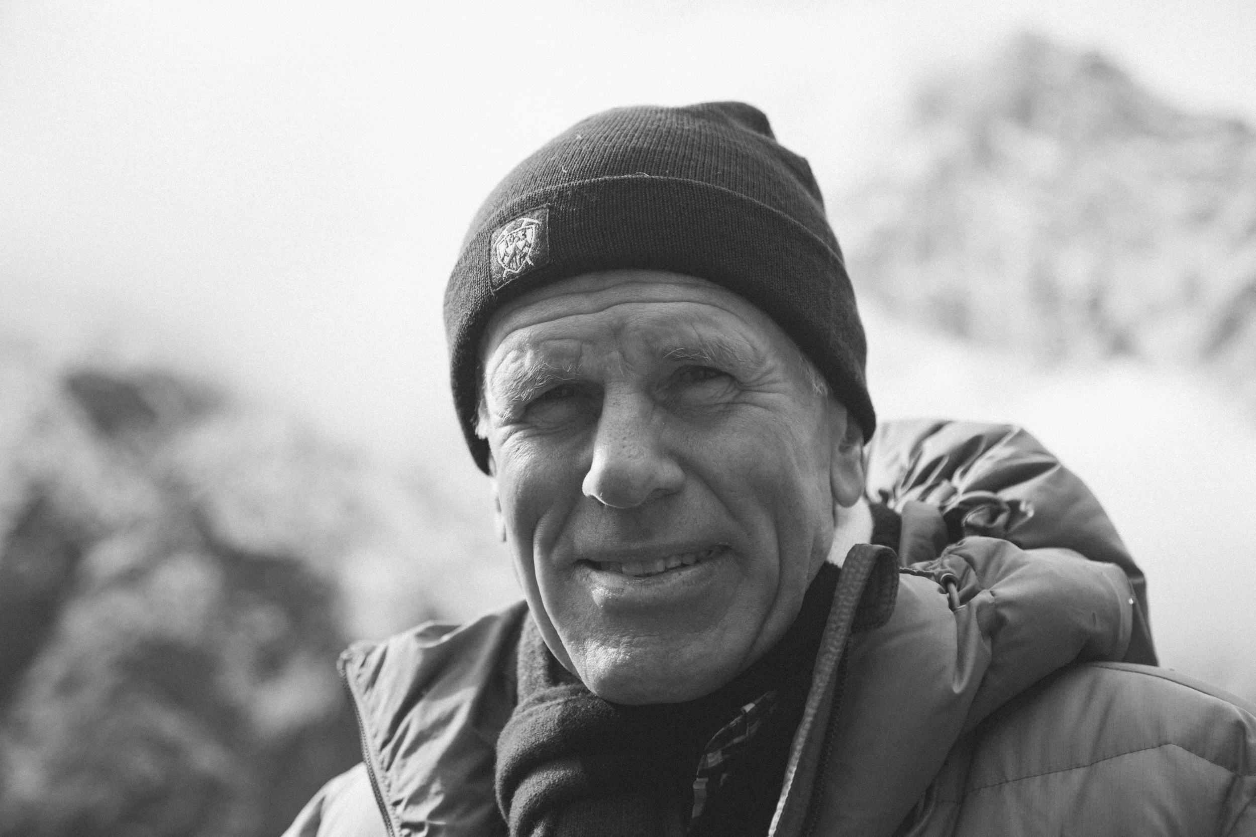 Black and white photo of an older man outdoors wearing a beanie hat and winter jacket, with a blurred mountain landscape in the background.