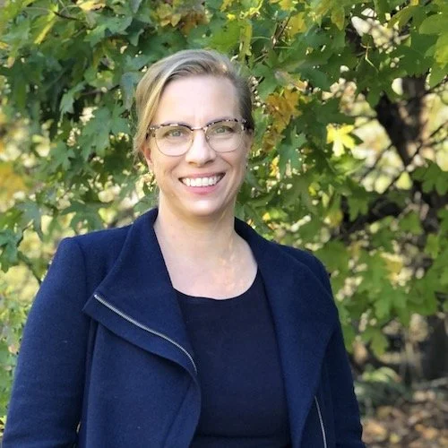 A woman with short blonde hair and glasses smiling outdoors in front of green leafy trees, wearing a navy blue jacket over a black top.