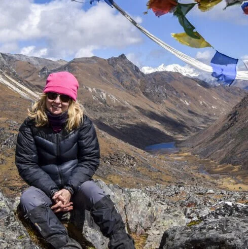 A woman sitting on rocks in a mountainous landscape with snowy peaks and a lake in the distance, wearing a pink hat, sunglasses, black jacket, and gray pants.