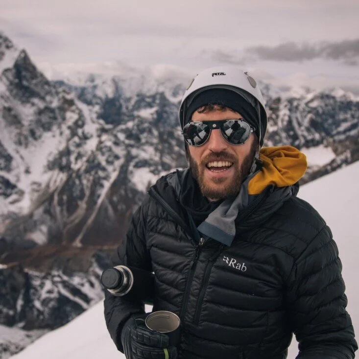 George Hillary dressed in black outdoor gear, wearing sunglasses and a helmet, stands on a snowy mountain with rugged, snow-covered peaks in the background.