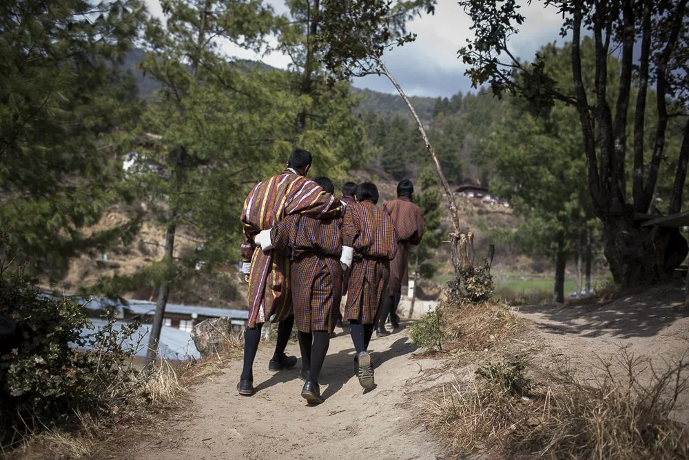 Group of Bhutan students in traditional school uniforms walking on a dirt path in a mountainous rural area surrounded by trees.