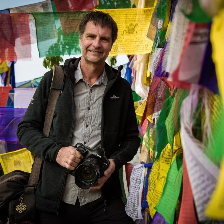 Richard I'anson smiling while holding a camera in front of colourful prayer flags.