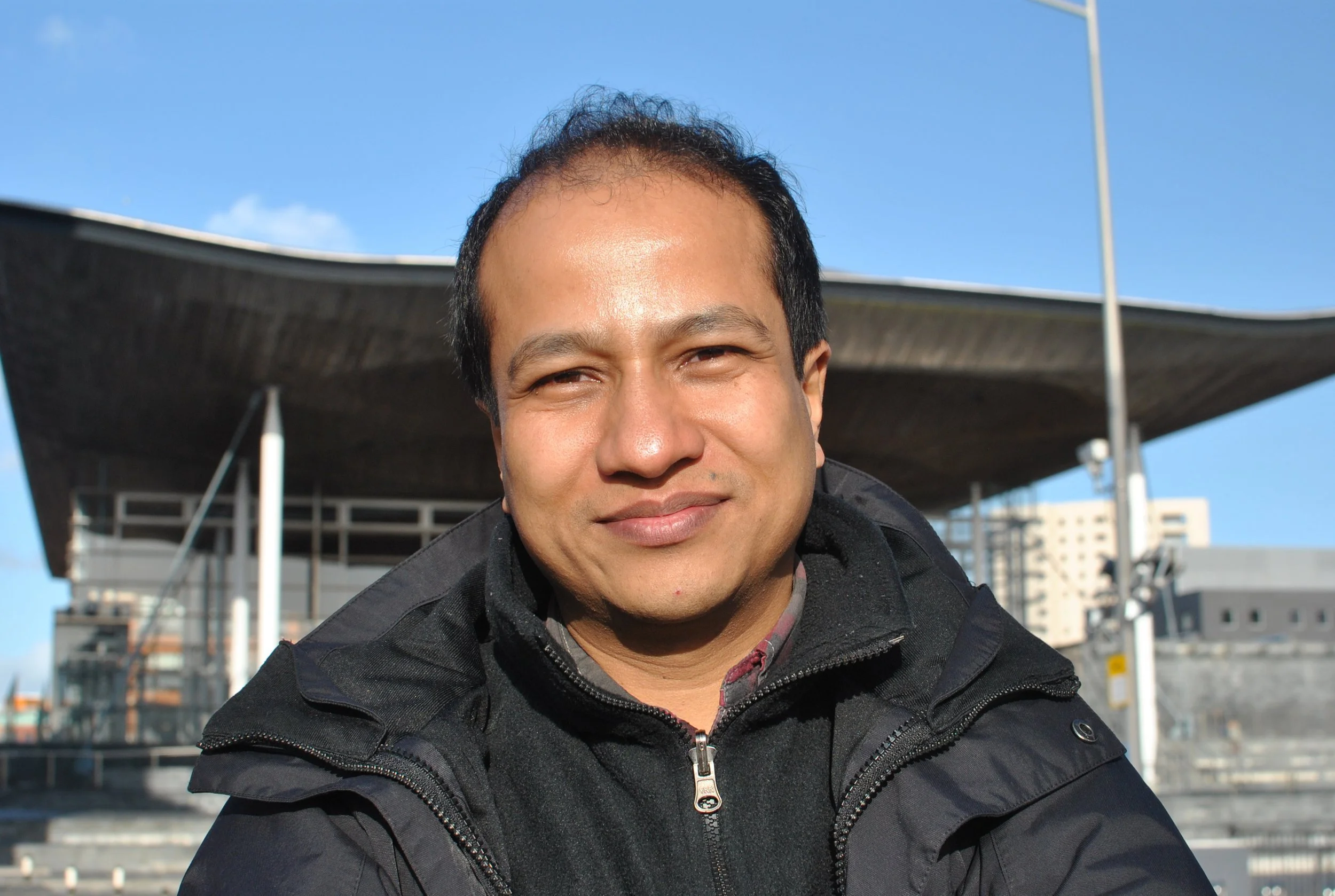 A man smiling outdoors on a sunny day, wearing a black jacket, with a modern building in the background.