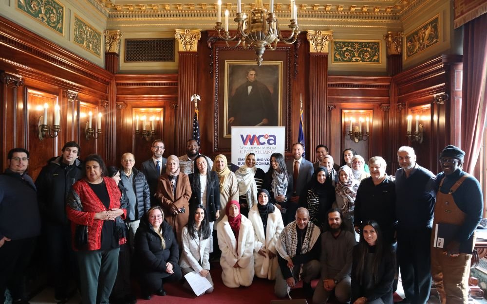 Group of diverse people posing inside a historic wood-paneled room with a large portrait of Abraham Lincoln, chandeliers, and a WCA (Wisconsin Muslim Civic Alliance) banner.