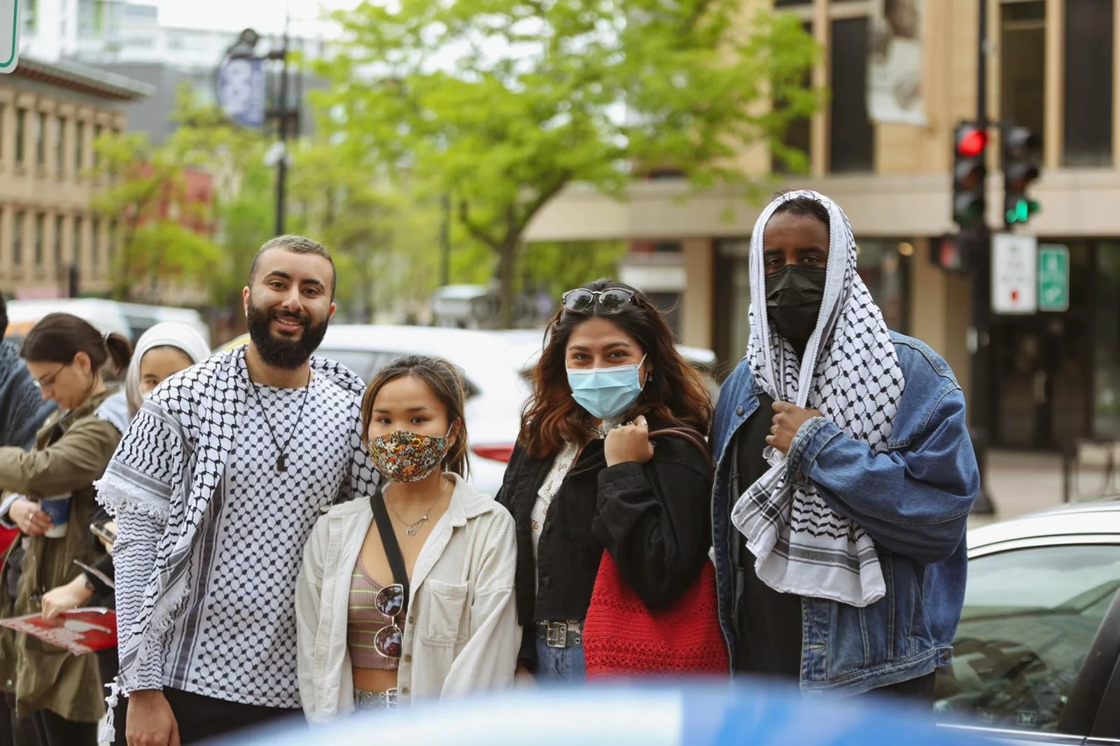 Group of four diverse people standing outdoors in a city street, all wearing face masks, with trees and buildings in the background.