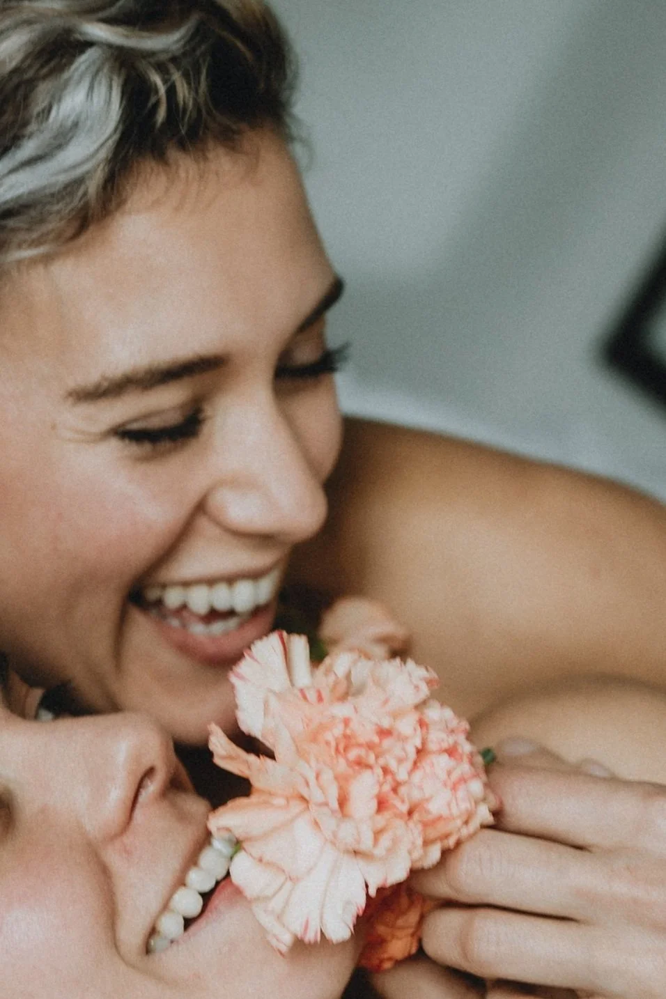 A woman smiling as she holds a pink flower near her face. Another person is close to her, partially visible.