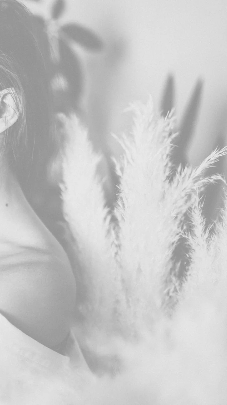 A person with long hair partially visible, standing behind tall, fluffy pampas grass in a blurred, grayscale setting.