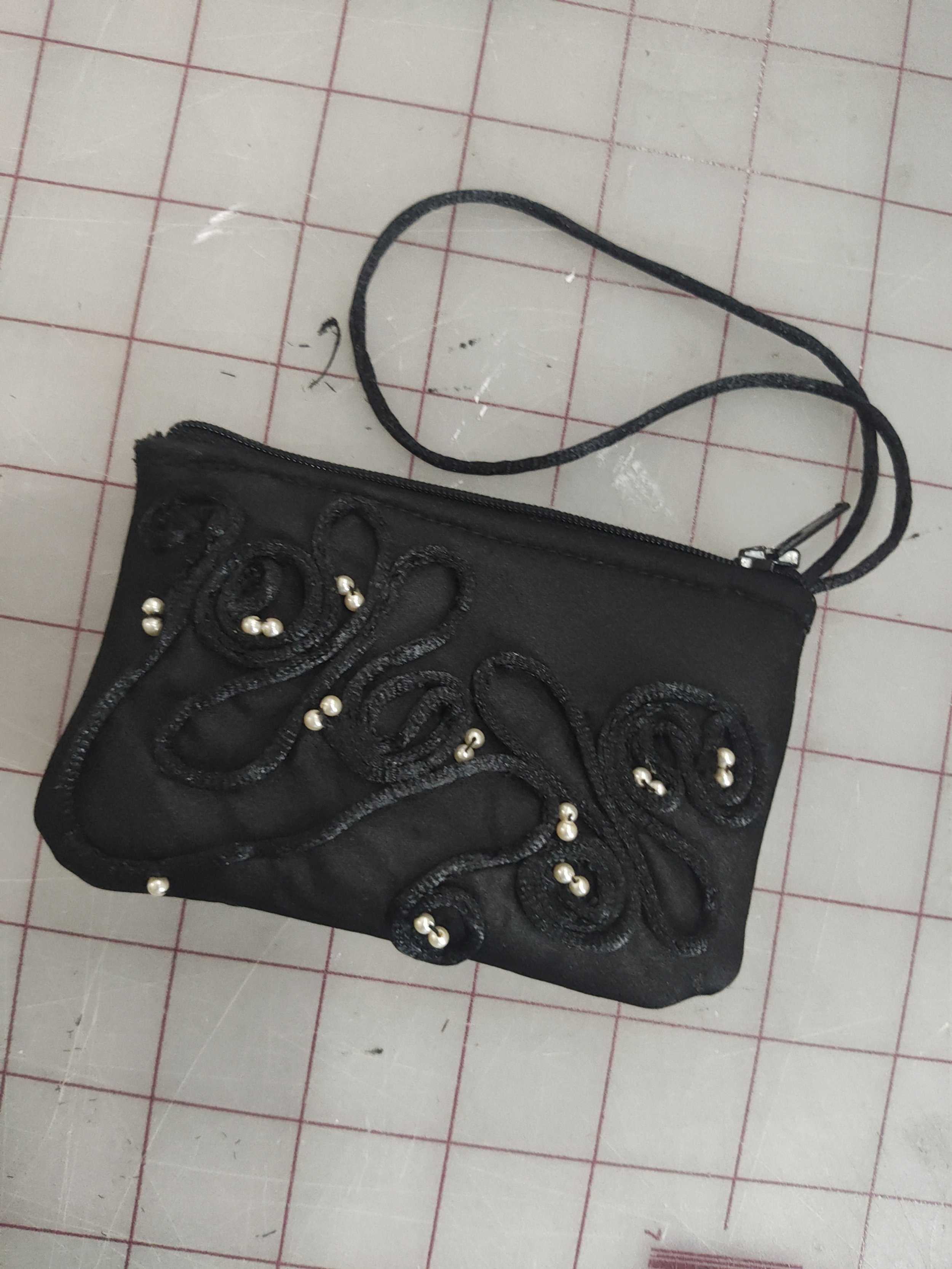 Black clutch purse with decorative embroidery and pearl accents, placed on a grid-lined surface.