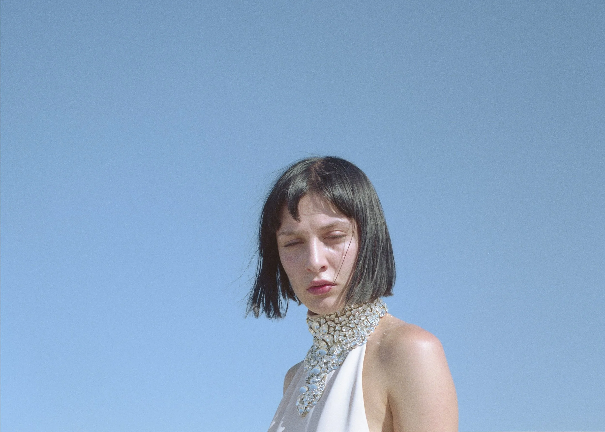 model with short black hair and fair skin poses in jewelled high neck dress against light blue sky