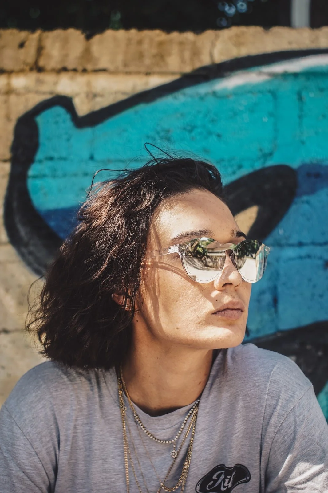 Model in grey t-shirt gold jewellery and sunglasses in front of graffiti 