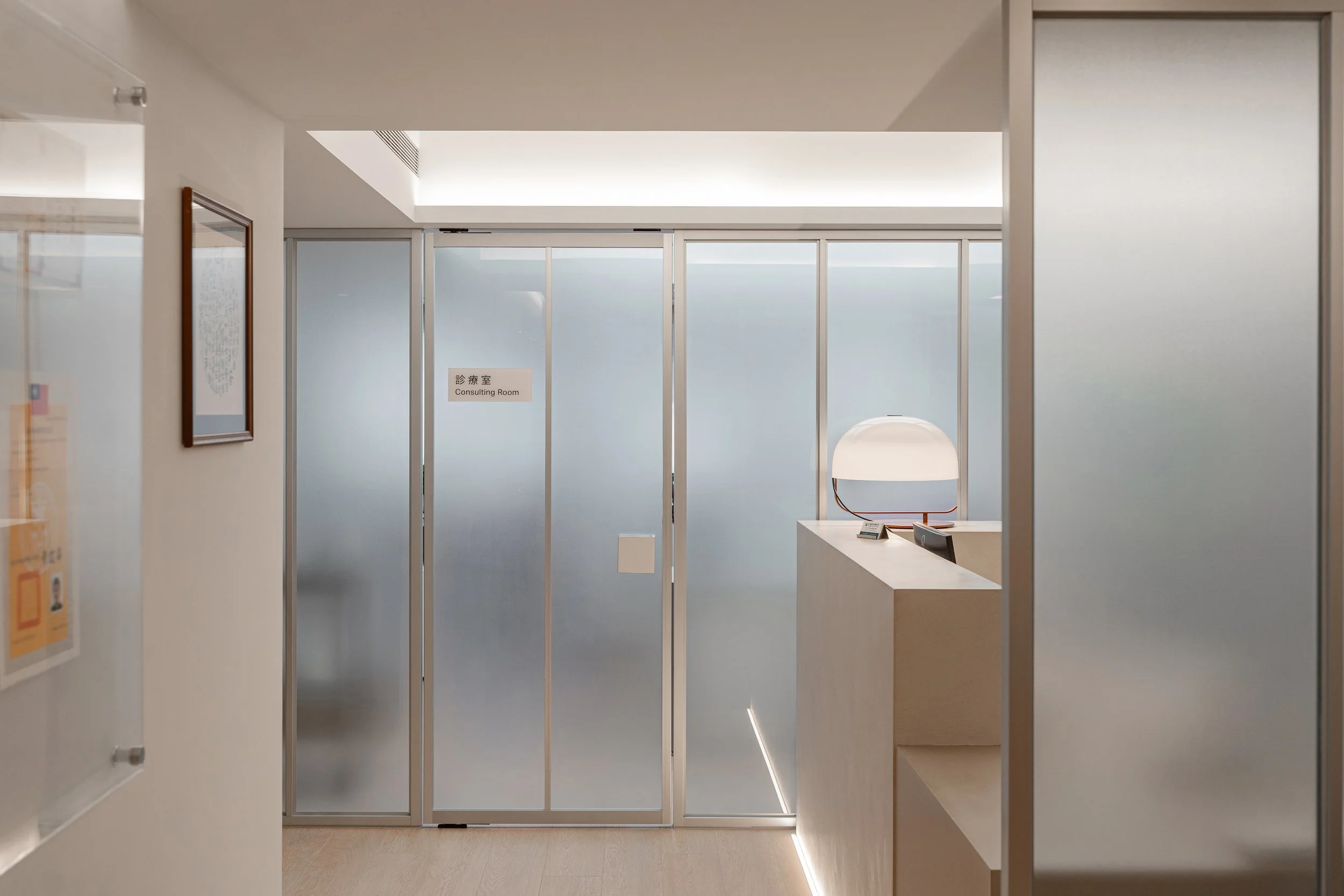 Reception area outside consultation room with frosted glass walls and a beige desk with a lamp, designed for privacy and a calm atmosphere.