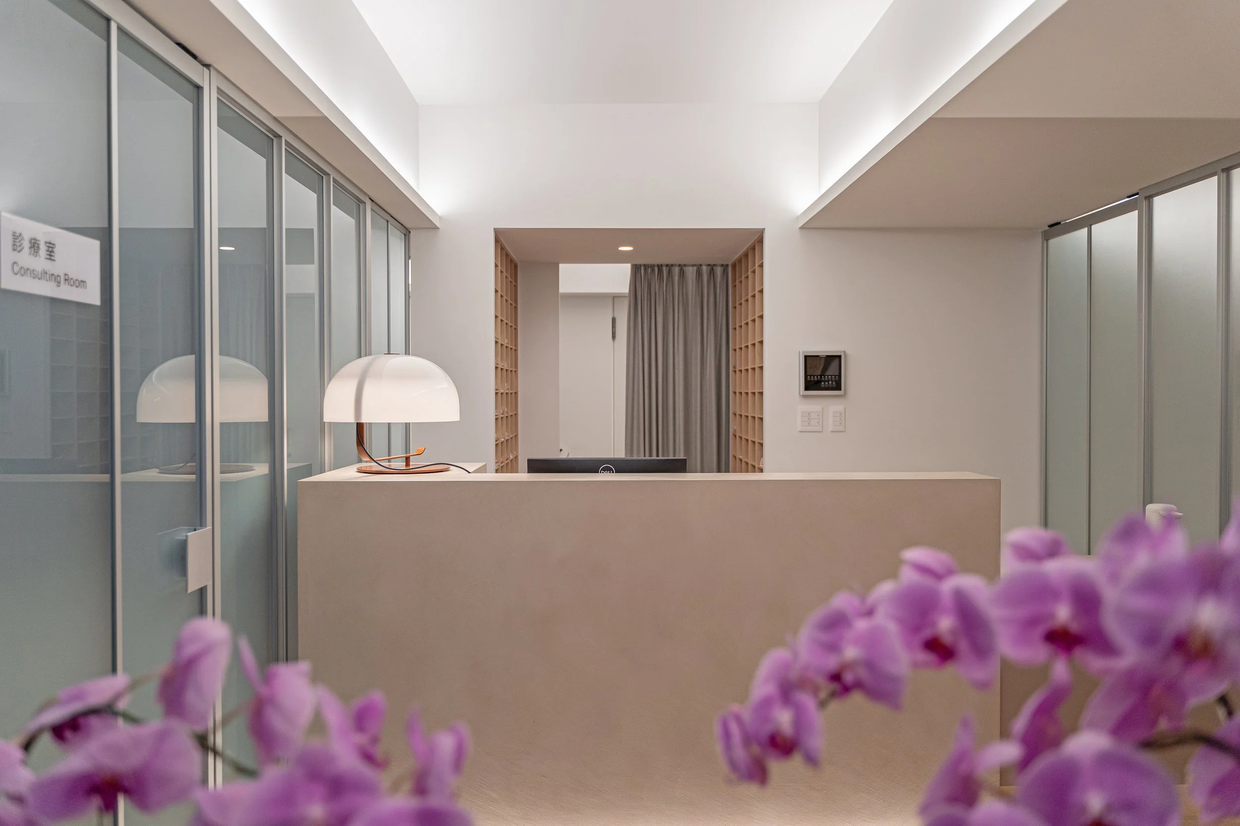 Modern clinic reception area with a beige front desk, white table lamp, glass partitions, and purple orchids adding a welcoming touch.