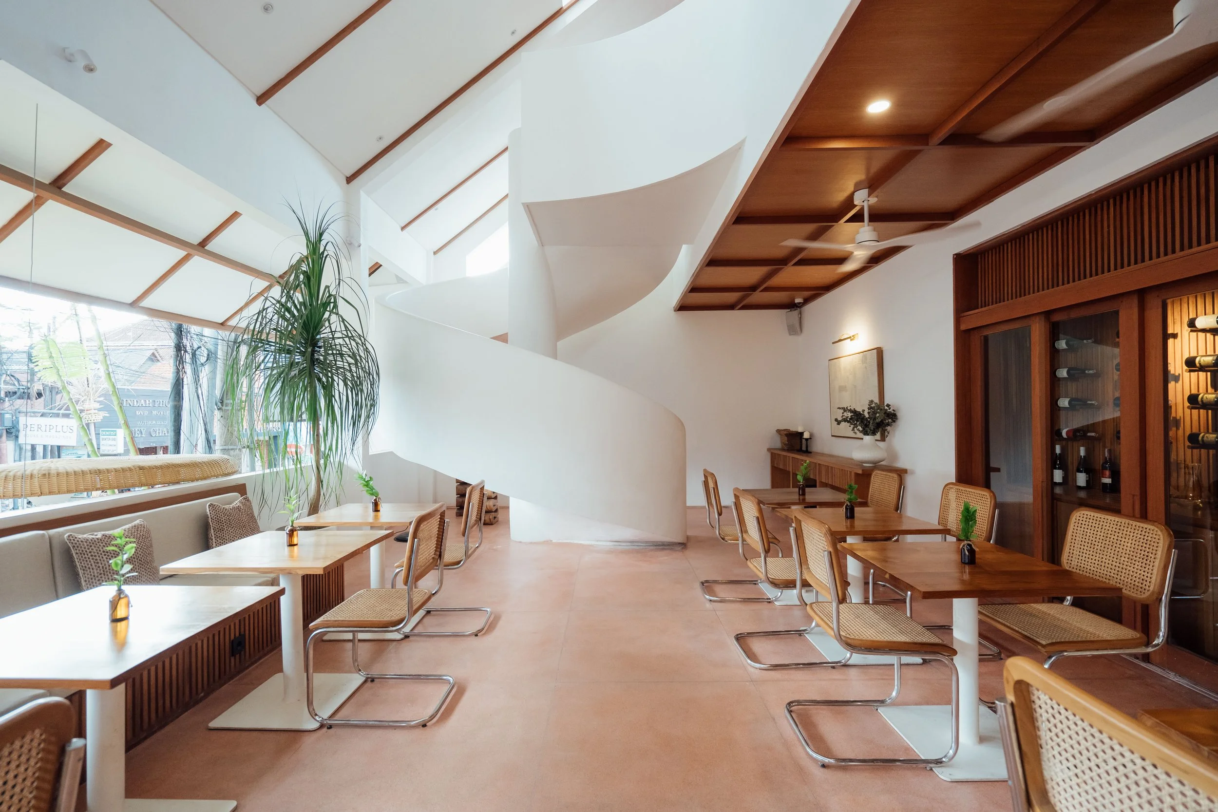 A cozy restaurant interior with natural lighting, wooden ceiling panels, white walls, and a mix of wooden tables and chairs. There is a large window with a view of the street outside, some indoor plants, a wine rack, and wall art. Ceiling fans and recessed lighting enhance the ambiance.