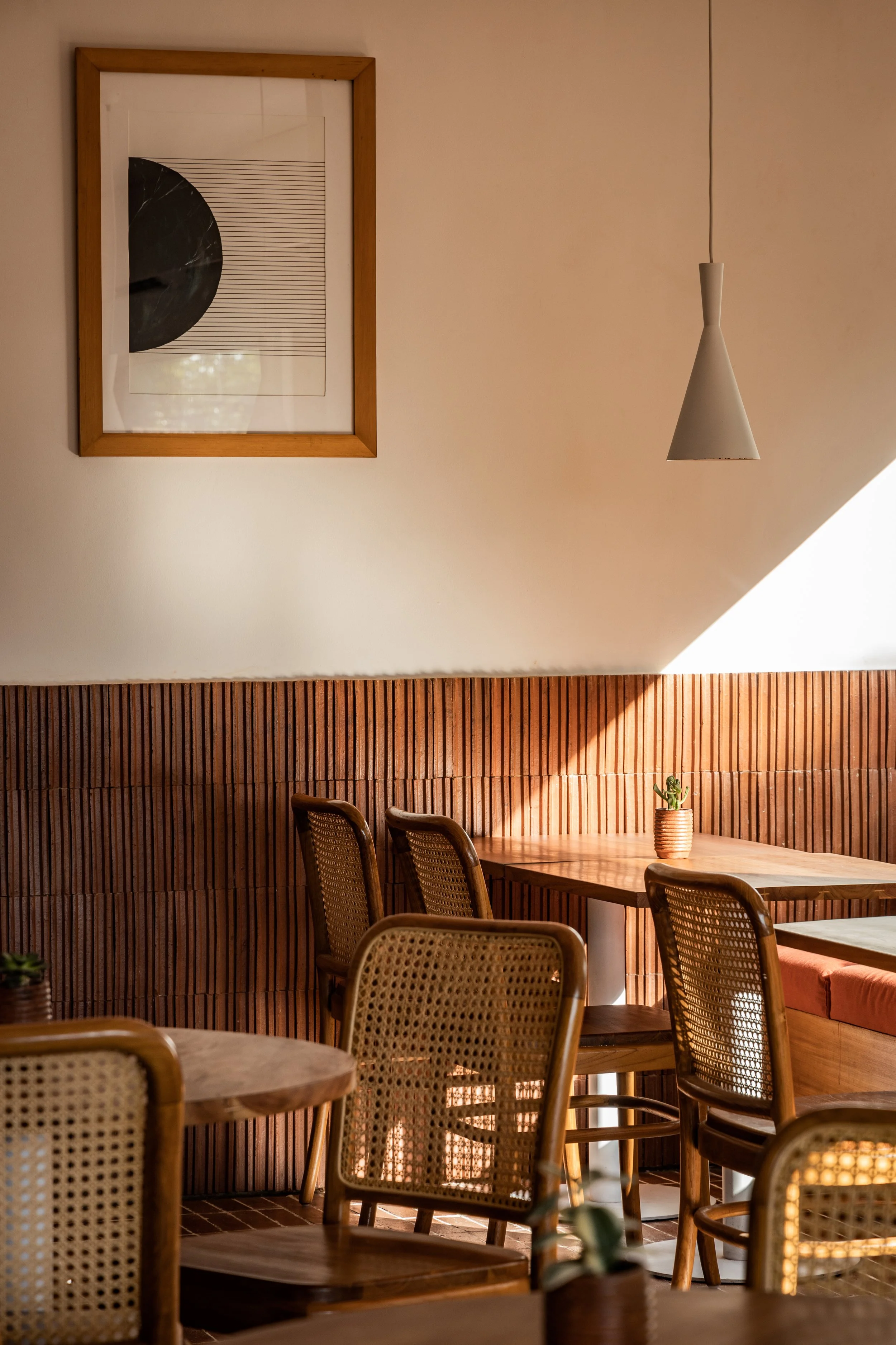 A cozy restaurant interior with wooden chairs and tables, a brown wood-paneled wall, a small potted plant on the table, modern wall art, and a cone-shaped pendant light hanging from the ceiling.
