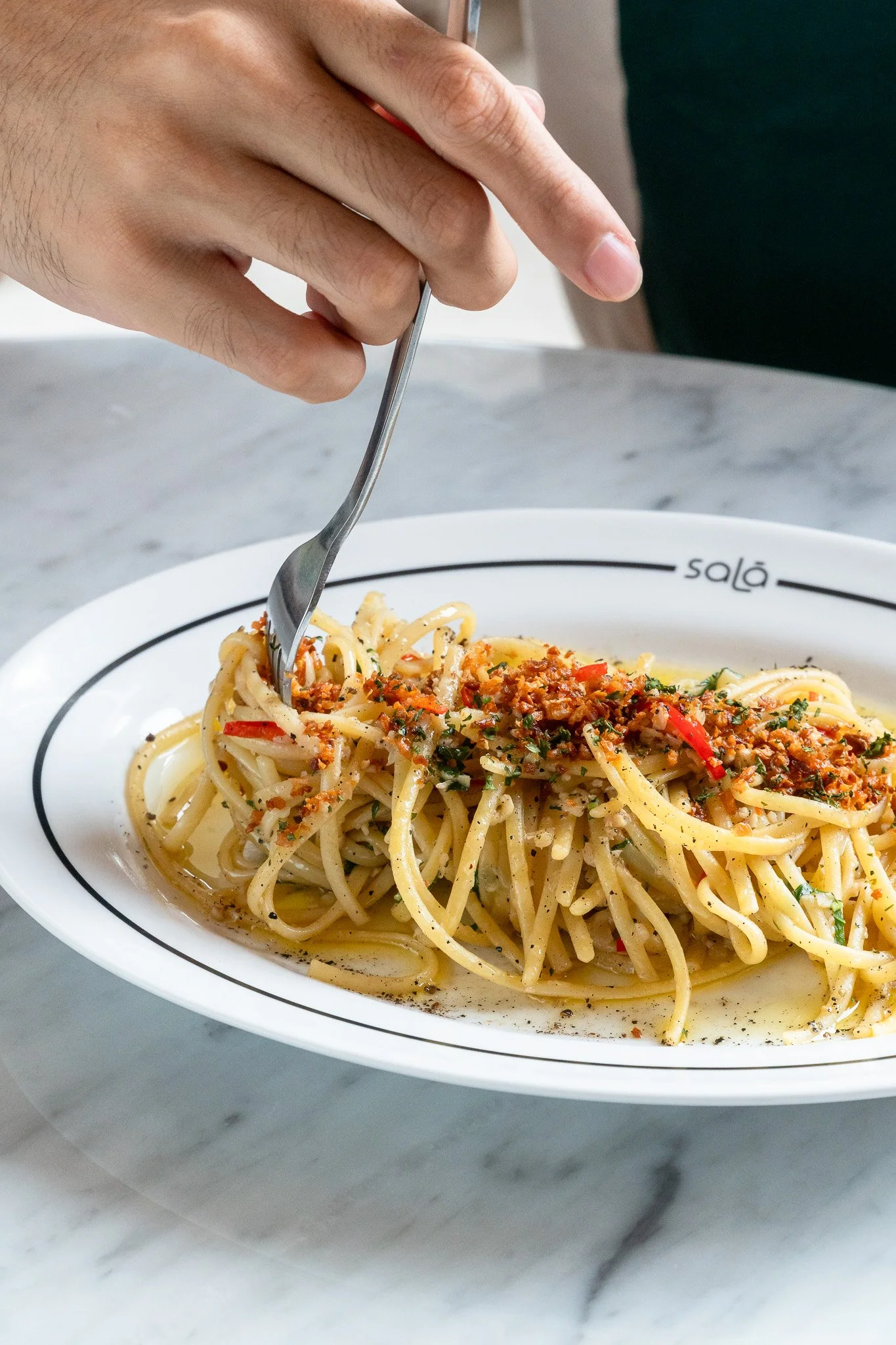 Close-up of a person using a fork to pick up a bite of spaghetti with tomato sauce and herbs from a white bowl on a marble surface.