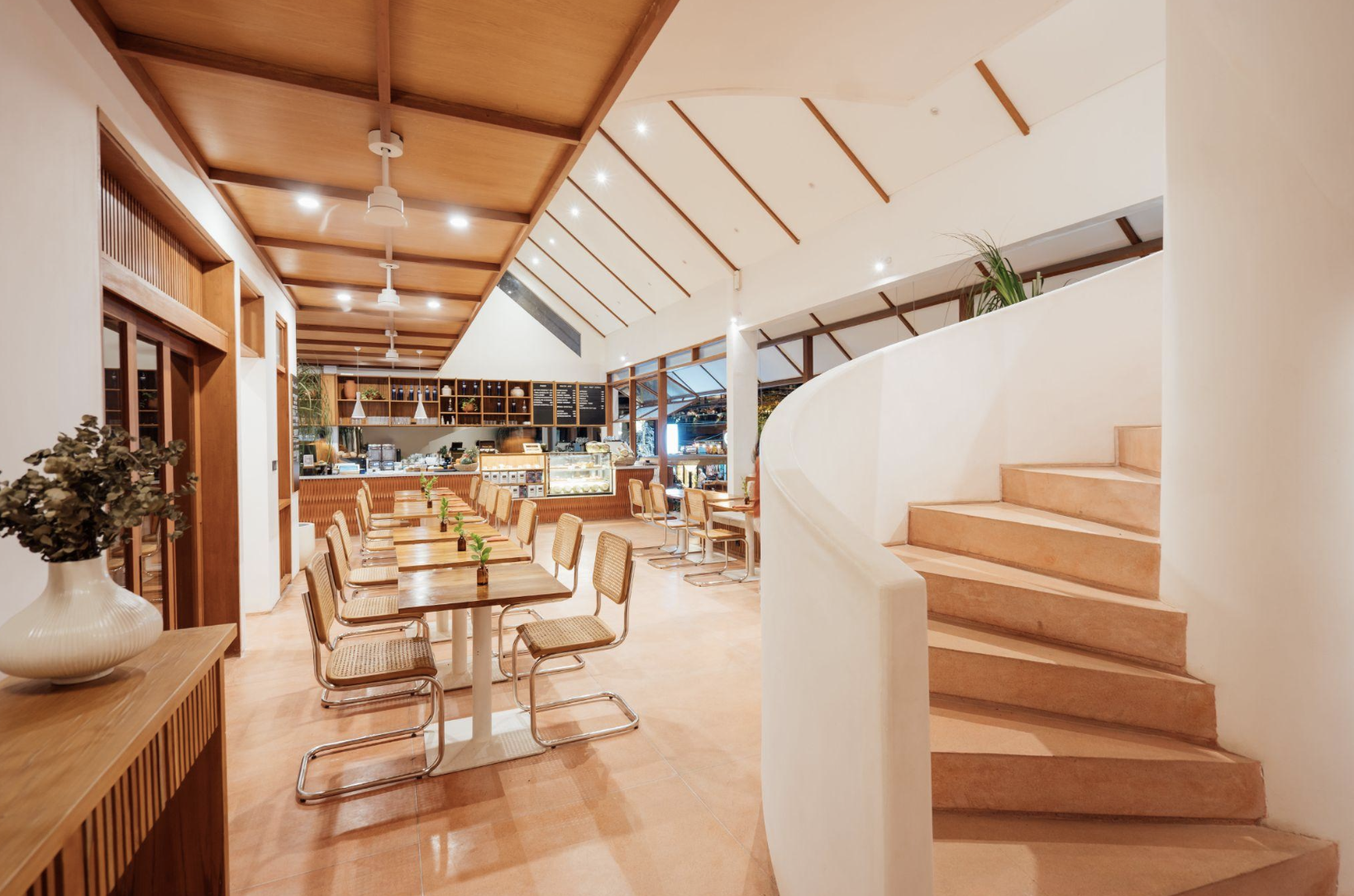 Interior of a modern cozy cafe with wooden accents, a staircase on the right, and a seating area with tables and chairs, a counter with menu boards, and potted plants.