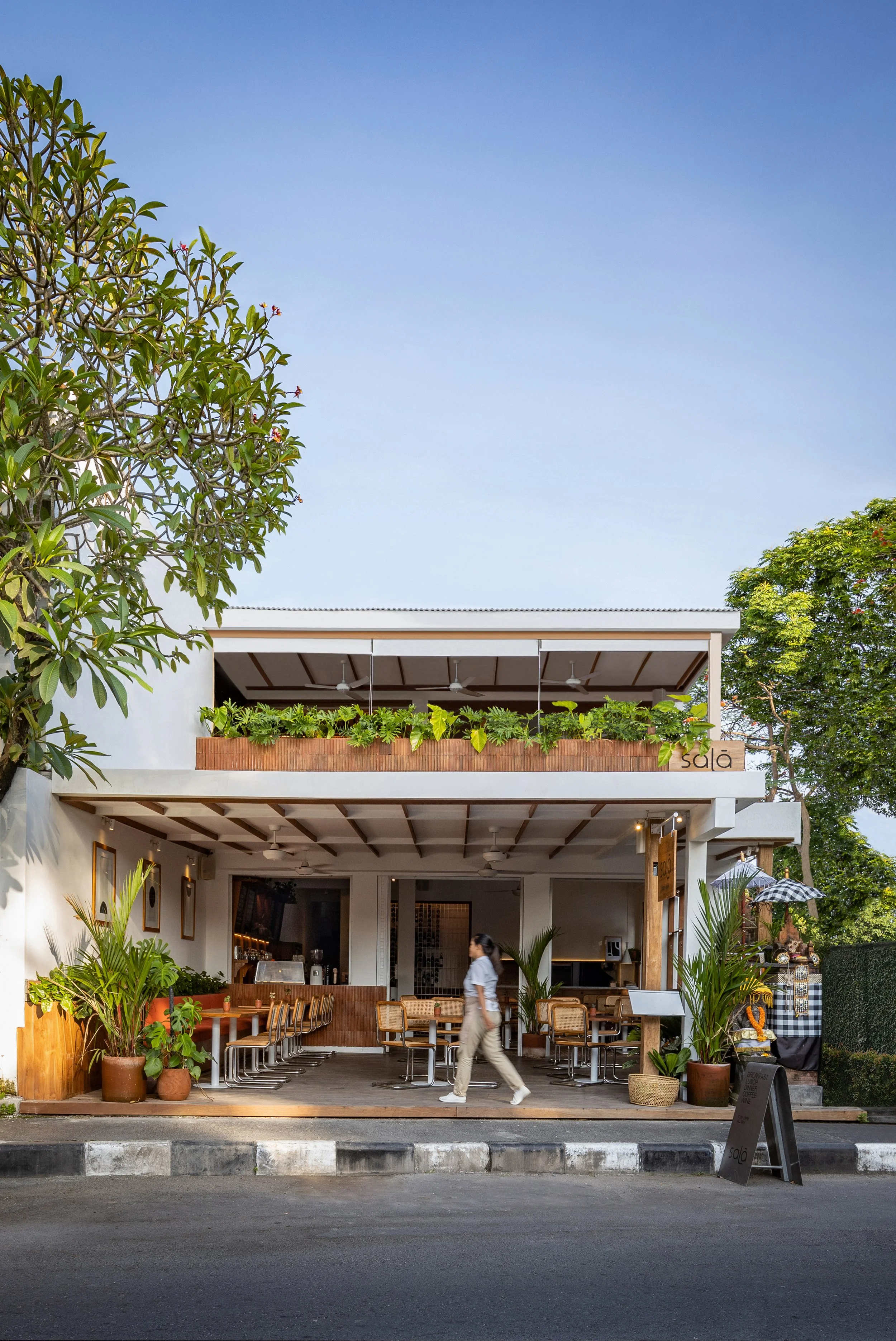 A two-story modern restaurant with outdoor seating on the ground floor, decorated with potted plants. The upper floor features a balcony with greenery and a sign that reads "sala." There are trees around the building and a person walking by in the foreground.
