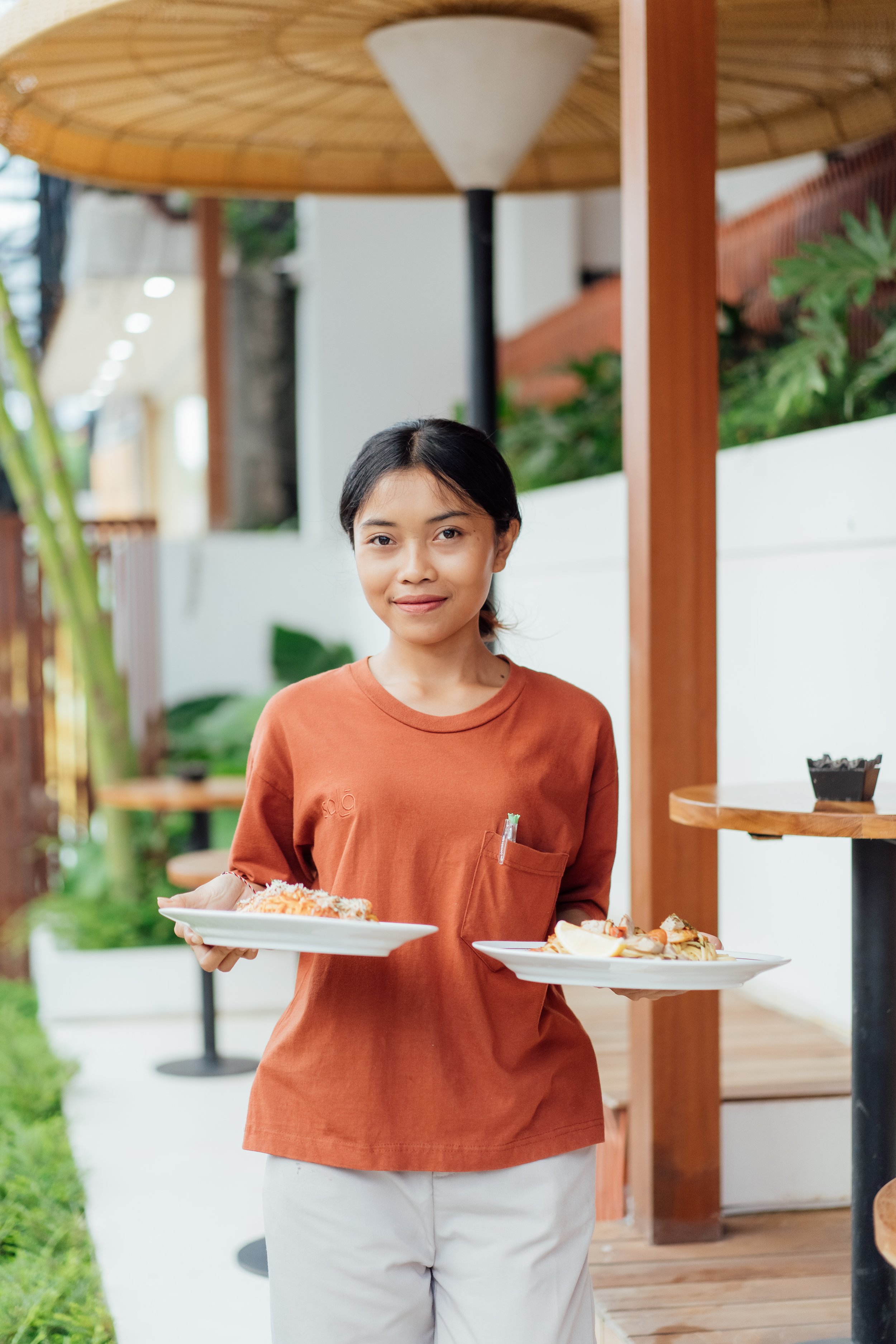 A young woman with dark hair, wearing an orange-brown shirt and white pants, standing outdoors in front of a white wall and holding two plates of food, smiling at the camera.