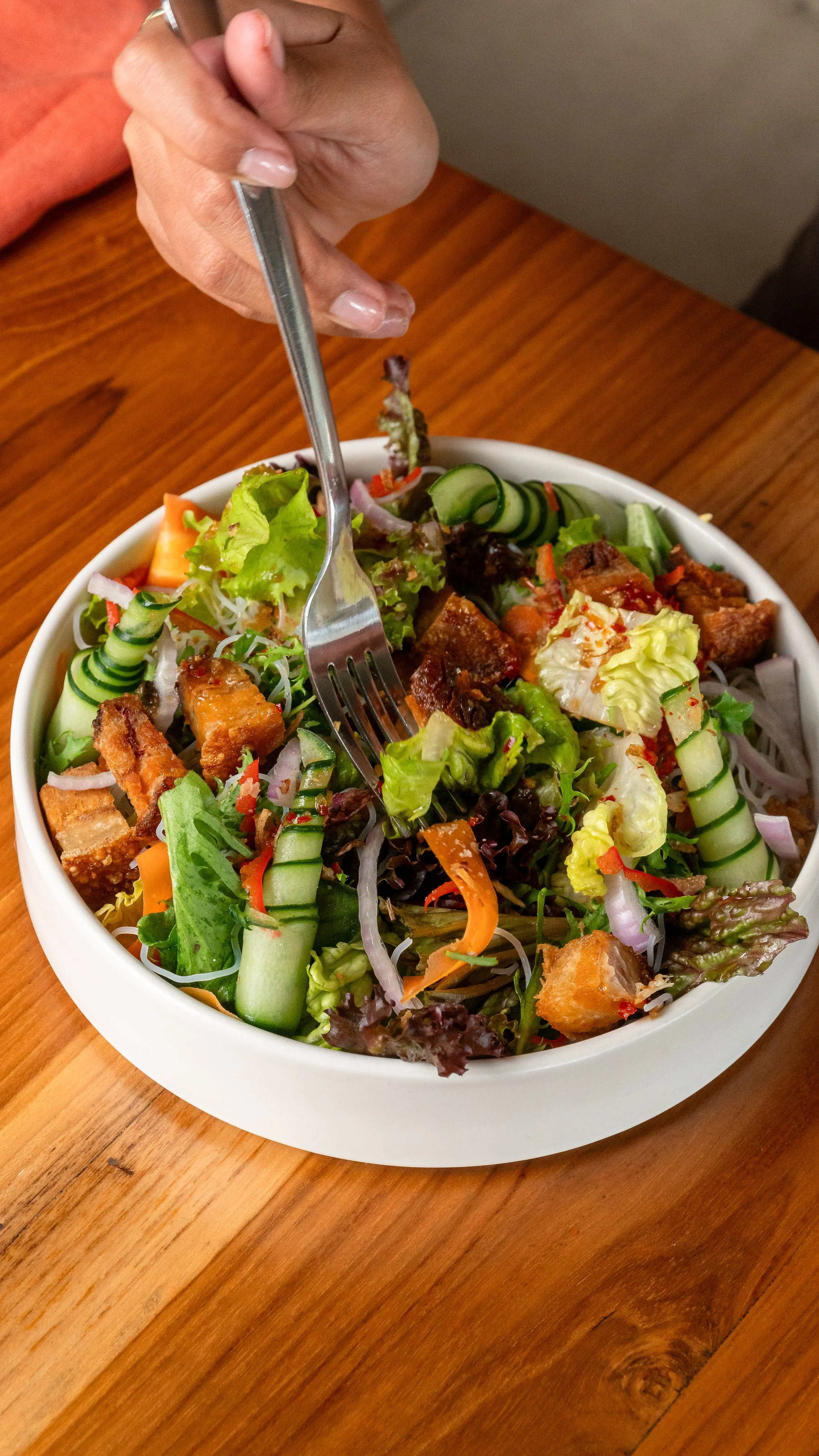 A person holding a fork over a colorful bowl of mixed salad with various greens and vegetables on a wooden table.