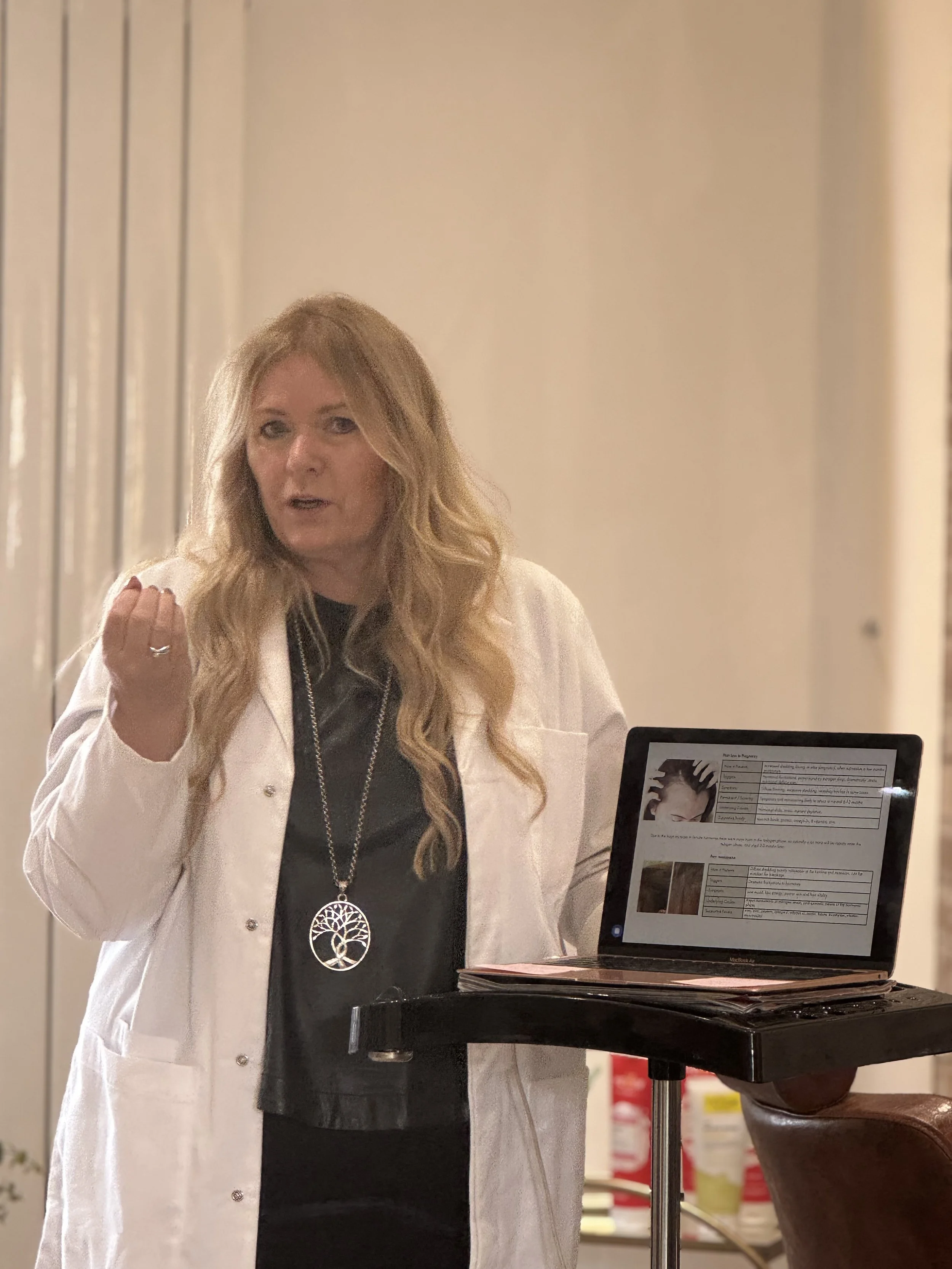 A woman with long wavy blonde hair, wearing a white coat and a necklace with a circular pendant, speaking or explaining something. She's standing behind a lectern with a laptop displaying notes or a presentation slide.