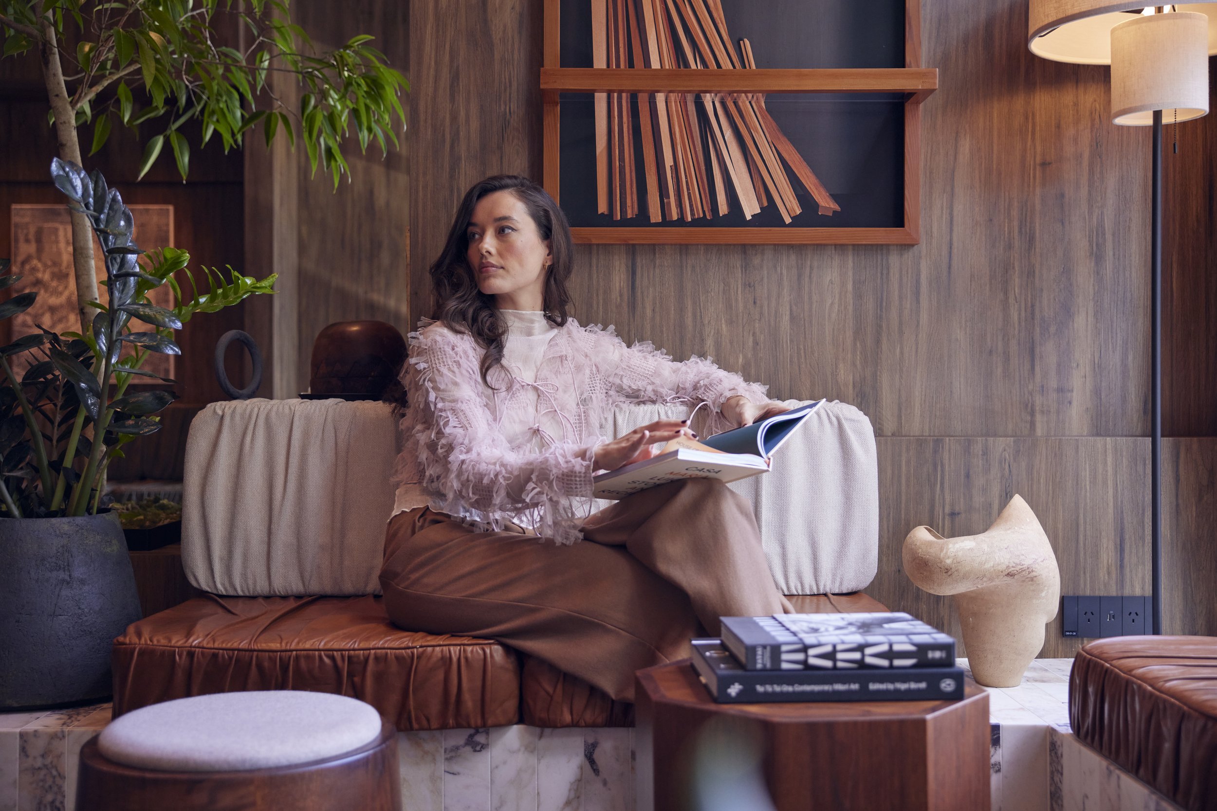 Woman sitting on a beige couch in a cozy, modern living room with wooden walls, green plants, and contemporary decor, reading a book.