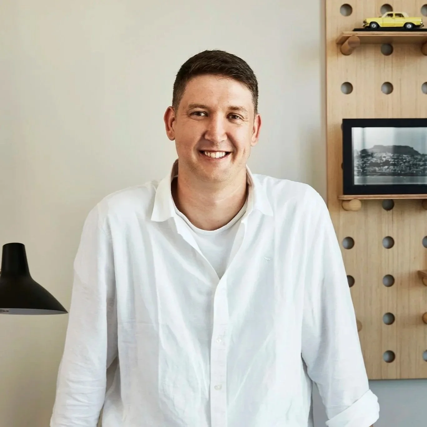 A smiling man with short dark hair wearing a white shirt, sitting at a desk with a black desk lamp and a wooden wall with a shelf and a framed black and white photo in the background.
