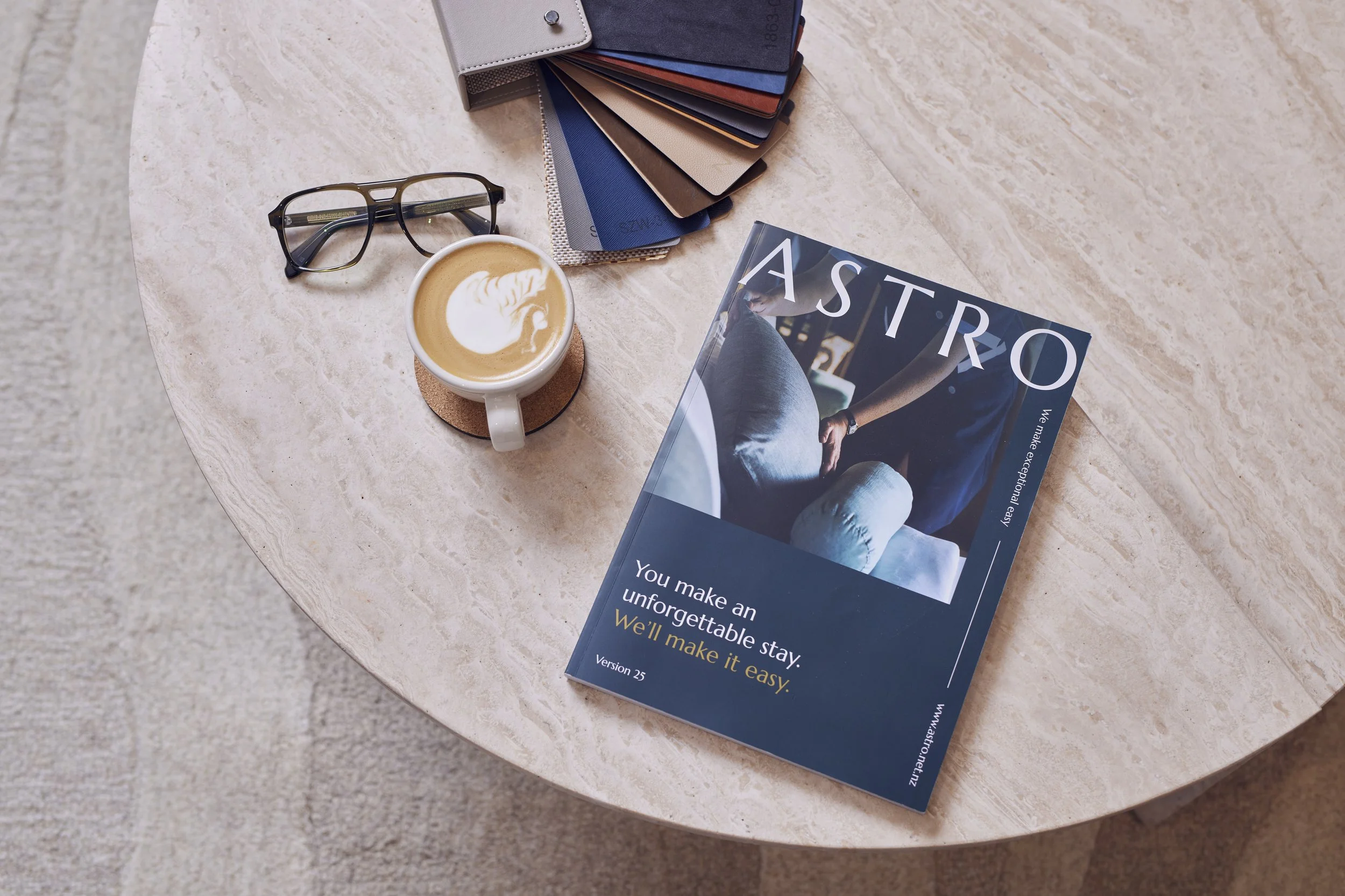 A marble round table with a color swatch sample book, a pair of glasses, a coffee cup with latte art, and a magazine titled 'ASTRO' on it.