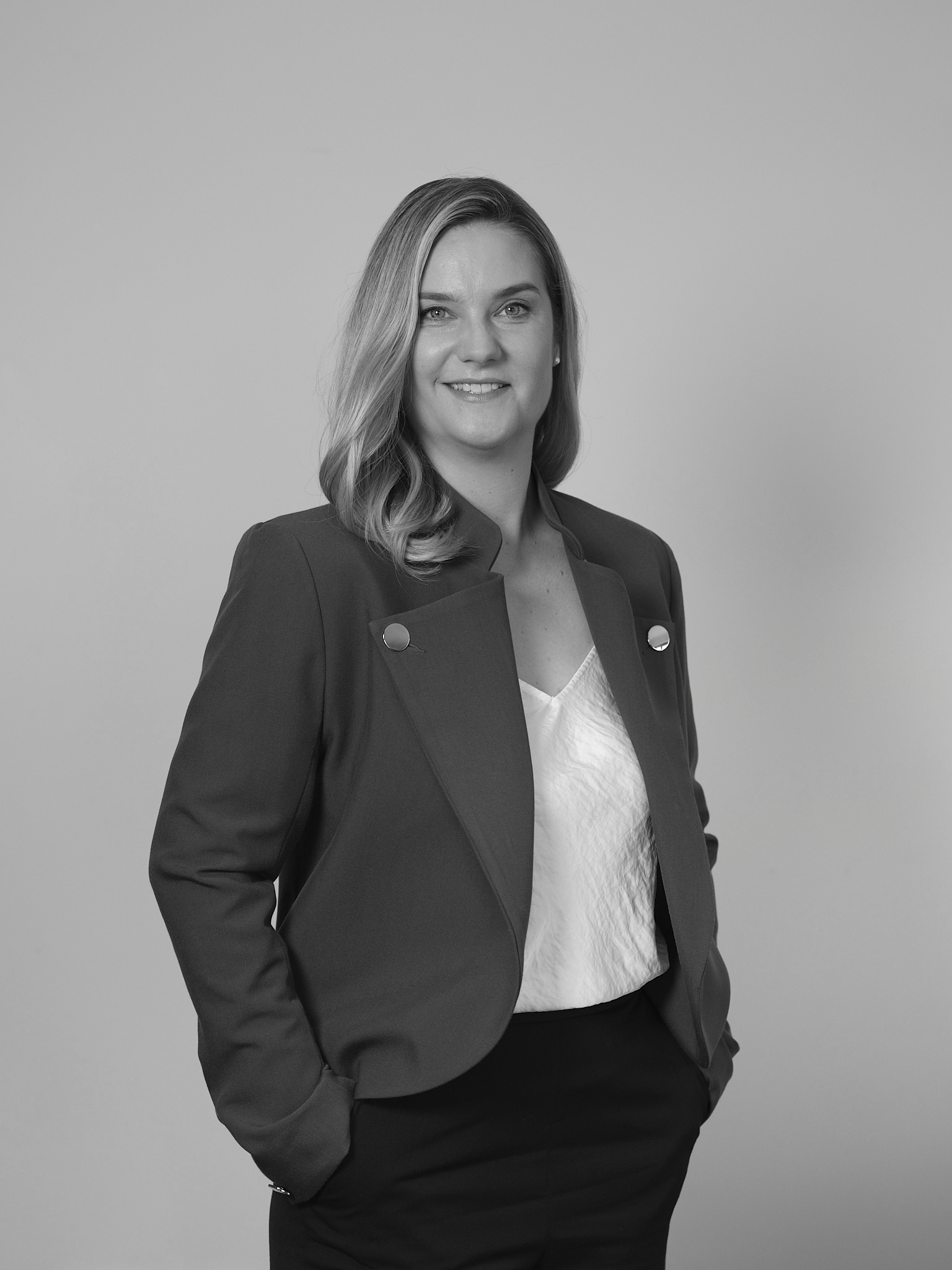 A black and white portrait of a woman with shoulder-length wavy hair, smiling, wearing a dark blazer with metallic buttons over a white top, standing against a plain background.