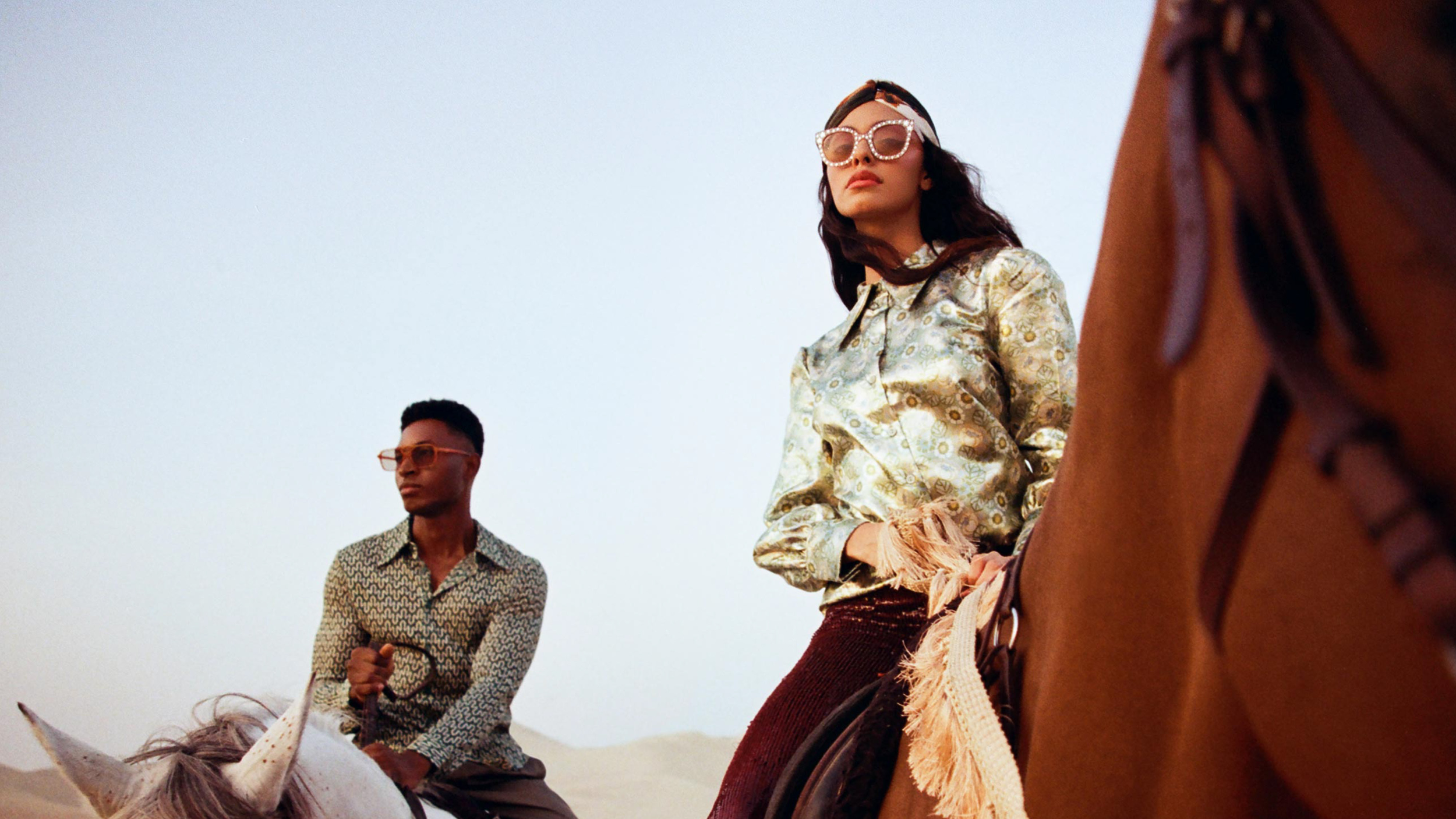 Two people on horseback in a desert or sandy area, with one woman wearing large sunglasses and vintage-style clothing, and a man in the background with glasses and a patterned shirt.