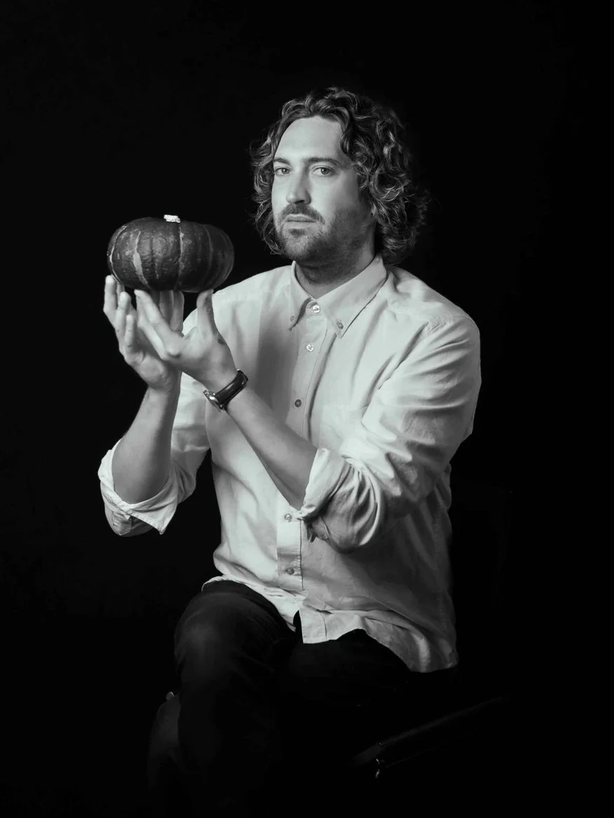 A man with curly hair and a beard holding a small pumpkin, dressed in a button-up shirt with rolled-up sleeves, sitting against a black background.