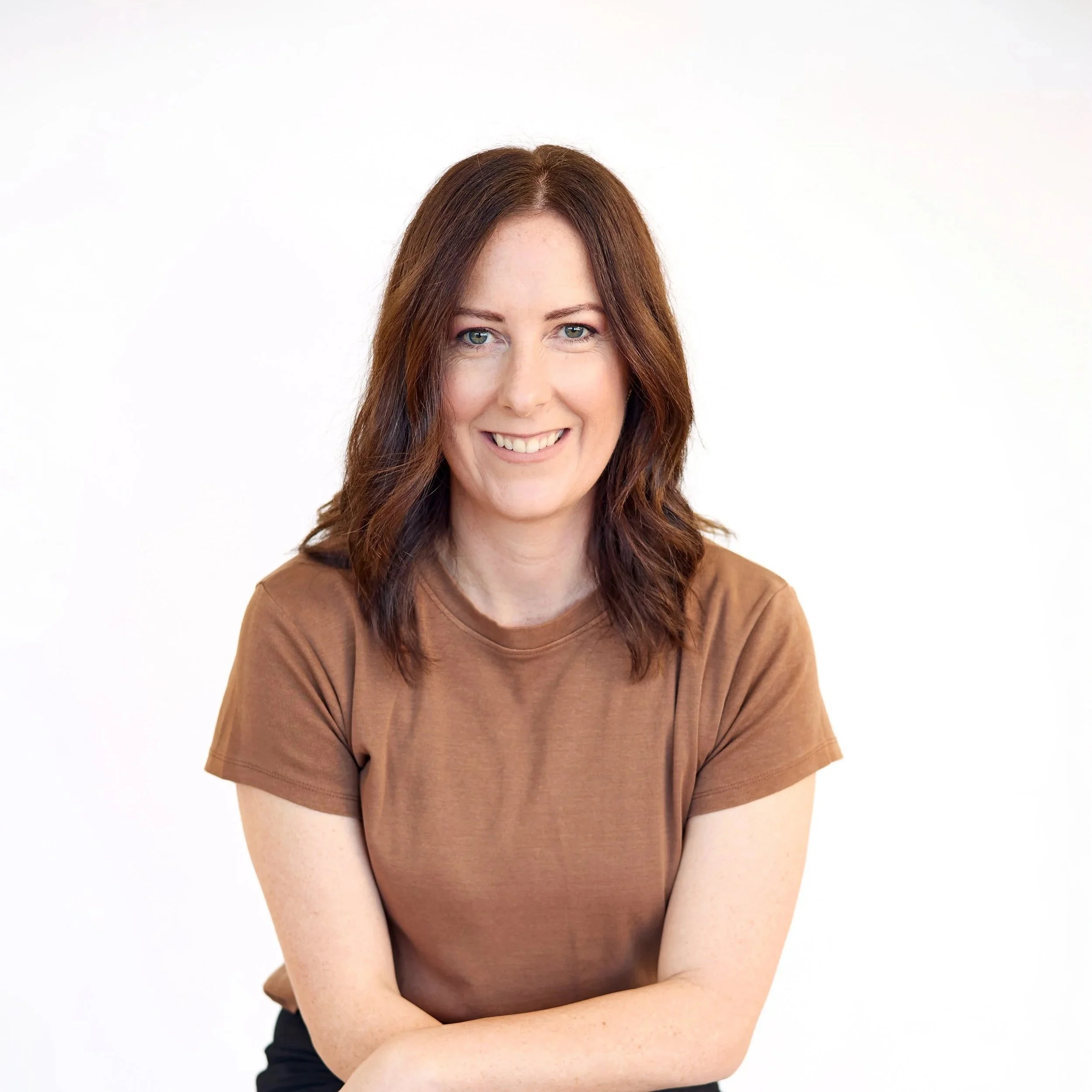 A woman with red hair and blue eyes smiling, wearing a brown short-sleeved top, with arms crossed in front of her against a plain light background.
