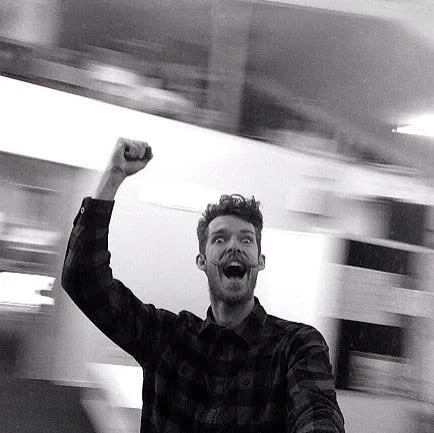 A man in a plaid shirt smiling and raising his fist in excitement, with a blurred background.