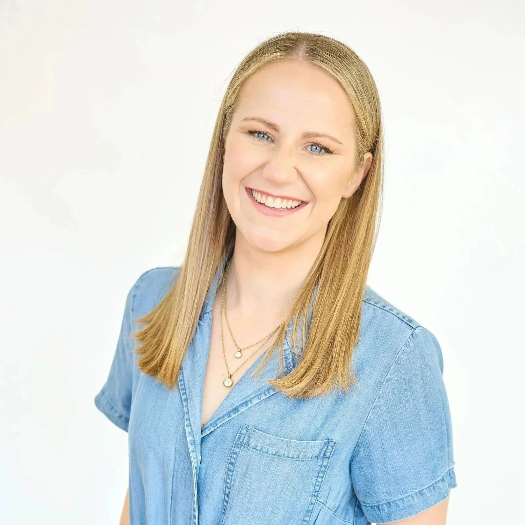 Close-up of a smiling young woman with long blonde hair, blue eyes, wearing a denim jacket and layered necklaces, against a plain white background.